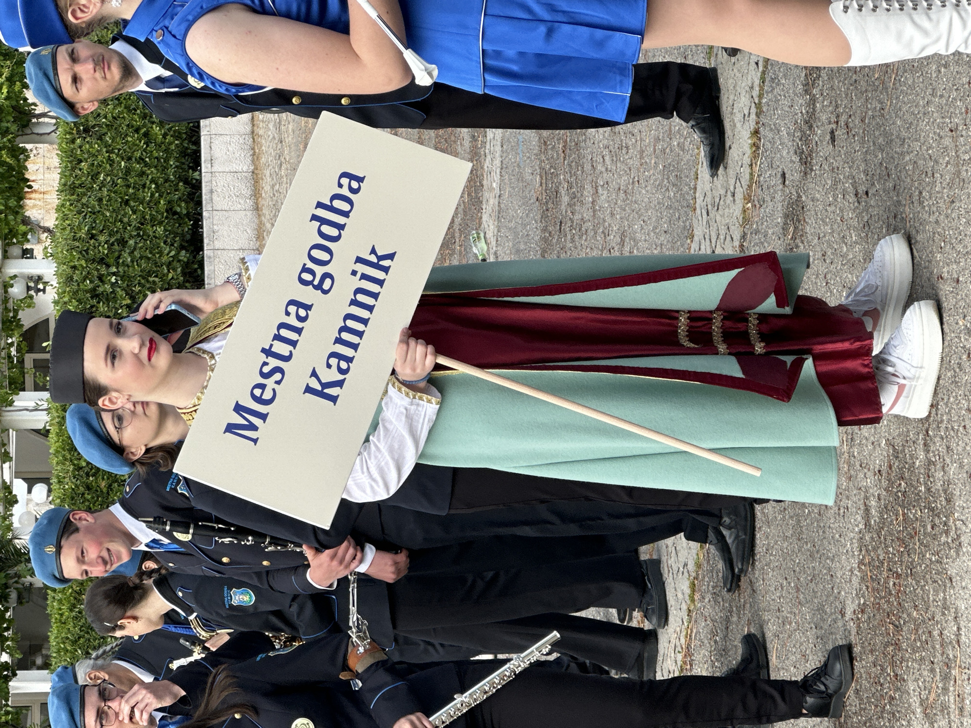 A long shot captures a marching band, facing right, on a paved outdoor street in Budva, Montenegro. The city is not visible in the background but is presumed based on the identification of the Mestna godba Kamnik banner. The group is mostly young, wearing a unified, formal uniform of black jackets with blue trim, black pants, black shoes, and a blue beret. They are playing musical instruments, primarily woodwinds like clarinets and flutes, and some carry batons. A few wear eyeglasses. One person carries a sign that says "Mestna godba Kamnik." A woman in traditional Slovenian clothing, including a black cap, white blouse, green skirt, and a decorative sash, stands in the middle of the group, holding the sign. She has fair skin, red lips, and gold ornamentation. She appears to be talking on a smartphone. Other uniformed members are visible both ahead and behind the sign carrier, continuing the line of the marching band. The ground is paved. The image is well-lit with natural sunlight. The shot is angled upward.