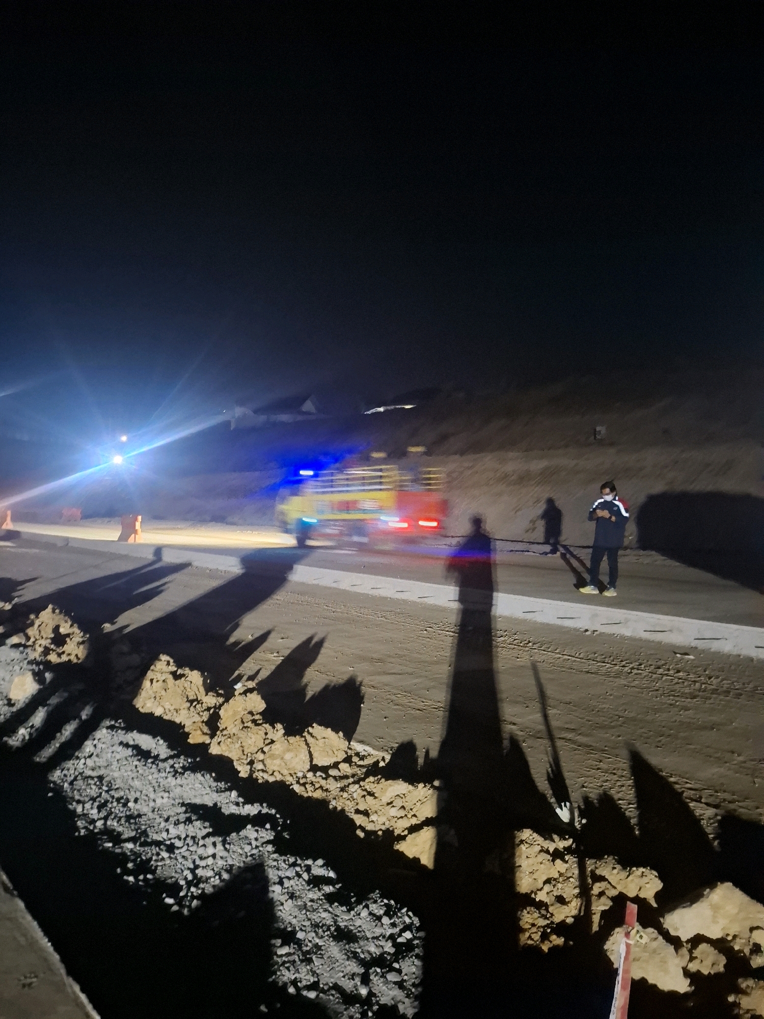 The image depicts a nighttime scene on a road in Karangtengah, Indonesia, possibly a construction site given the presence of soil mounds and a trench in the foreground. A blurry yellow truck with flashing red and blue lights, resembling an emergency vehicle, is moving on the road. In the middle ground, a person wearing a dark jacket with red and white accents and a face mask is standing and looking at their phone. Their shadow is cast on the ground. Further back, another blurry figure can be seen near the truck. The environment is dark, illuminated by artificial lights from the vehicle and potentially surrounding work lights, with a clear night sky above. Orange traffic cones are visible on the left side of the road. The ground is a mixture of dirt, gravel, and what appears to be a paved surface.