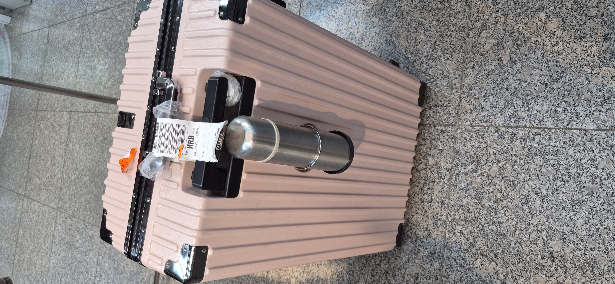 A light pink, ribbed suitcase is positioned upright on a speckled gray tiled floor. A metallic thermos is secured to the side of the suitcase with a ring holder. An airline baggage tag is attached to the suitcase handle, displaying text including "TO: HRB", "CA 1621 20MAR", and "VIA". The suitcase has black corner protectors and a black handle. The background shows more of the tiled floor and a metal pole. The scene is indoors, likely an airport terminal, given the baggage tag. There are no people visible in the image.