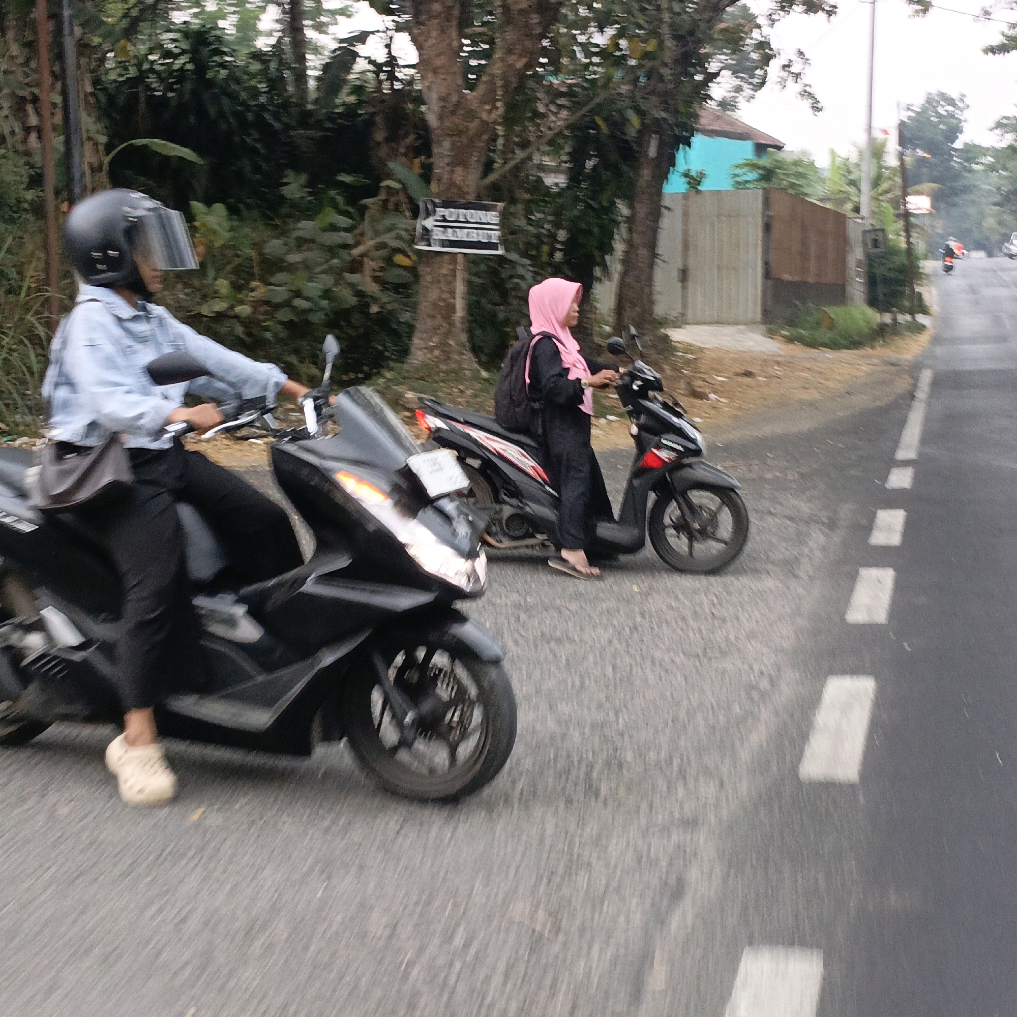 The image depicts two individuals riding motorcycles on a paved road in Salatiga, Indonesia. The foreground shows a person wearing a light blue denim jacket and a black helmet with a clear visor, riding a black motorcycle. They are wearing black pants and light-colored slip-on shoes. A dark brown bag is slung over their left shoulder. In the mid-ground, another person wearing a black outfit and a pink hijab is riding a black motorcycle. They are carrying a black backpack. The environment suggests an outdoor, possibly semi-urban or rural setting, with trees and vegetation visible along the side of the road. A signpost with text in Indonesian is partially visible on the left. The road has painted white dashed lines, indicating traffic lanes. The lighting suggests it is daytime, possibly overcast or early morning/late afternoon. The overall impression is of everyday transportation on a street in Salatiga.