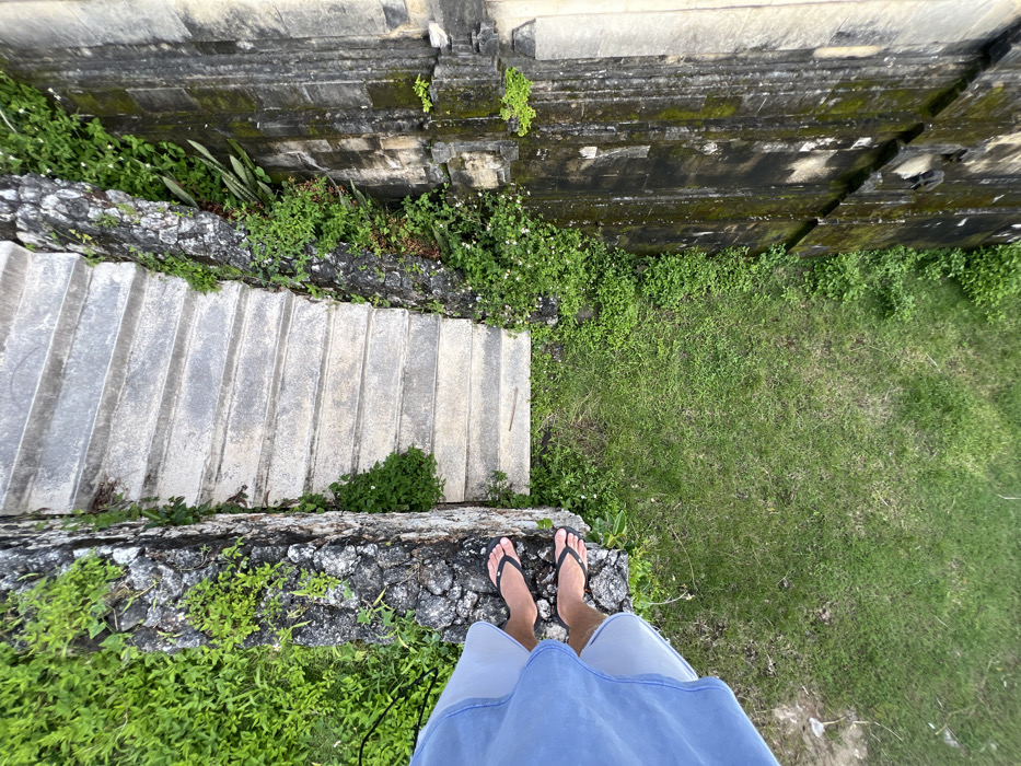 A high-angle forensic shot captures a view looking down into a stone stairwell in Pecatu, Indonesia. A person's bare feet, adorned with black flip-flops and partially obscured by light blue shorts, are positioned at the upper left edge of a rough-hewn stone wall. Below and extending diagonally from the upper center, a series of worn concrete steps descends between two vertical structures. The immediate left structure is a dark stone wall heavily covered with vibrant green foliage. The opposing right structure is a darker, possibly ancient or decorative carved stone wall, exhibiting weathering, discoloration, and mossy patches. A section of green grass is visible at the bottom of the stairwell, directly beneath the observer and to the right of the steps.
