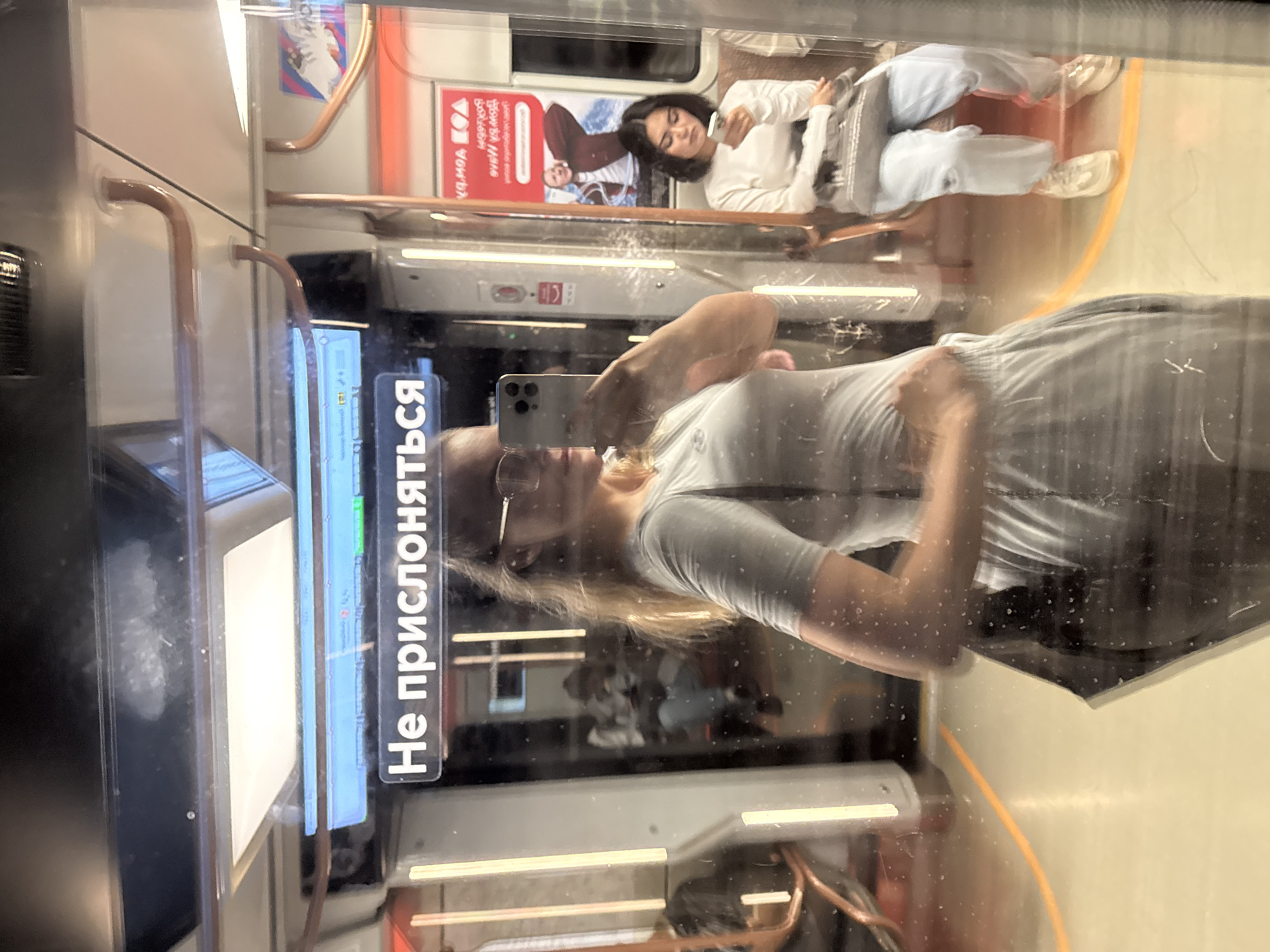 The image is a selfie taken inside a Moscow Metro carriage. A young woman with blonde hair and glasses is visible in the foreground, taking the photo with her smartphone. She is wearing a light grey t-shirt and grey sweatpants. To her right, reflected in the glass, another young woman with dark hair is seated, looking at her phone. She is wearing a white long-sleeved shirt and light blue pants. The carriage interior features bright vertical lights, brown handrails, and a sign in Russian reading "Не прислоняться" (Do not lean). Advertisements are visible on the walls of the carriage. The floor of the carriage is a light beige color with orange markings.