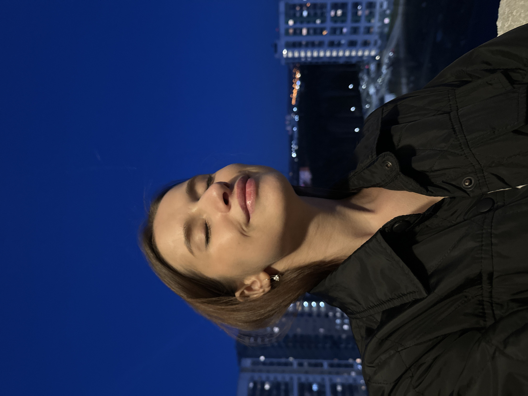 A young woman is depicted in a close-up, rotated portrait taken at night in an urban setting. Her head is tilted upwards, eyes closed, and lips slightly parted, conveying a serene or contemplative expression. She has light brown hair and wears a black, possibly puffer-style, jacket with snap buttons visible on the collar and sleeve. A small, shiny earring is visible on her right ear.
The background consists of a dark blue night sky, indicating a clear evening. Below the horizon, illuminated windows of multiple buildings create a blurred city skyline. These city lights are reflected as bright, elongated blurs on a dark surface in the lower part of the background, strongly suggesting the presence of a body of water, such as a river or canal. The image captures a quiet moment against the backdrop of a vibrant night city, possibly taken from a bridge or waterfront. No text is visible in the image. The location is contextualized as Yekaterinburg, Russia.