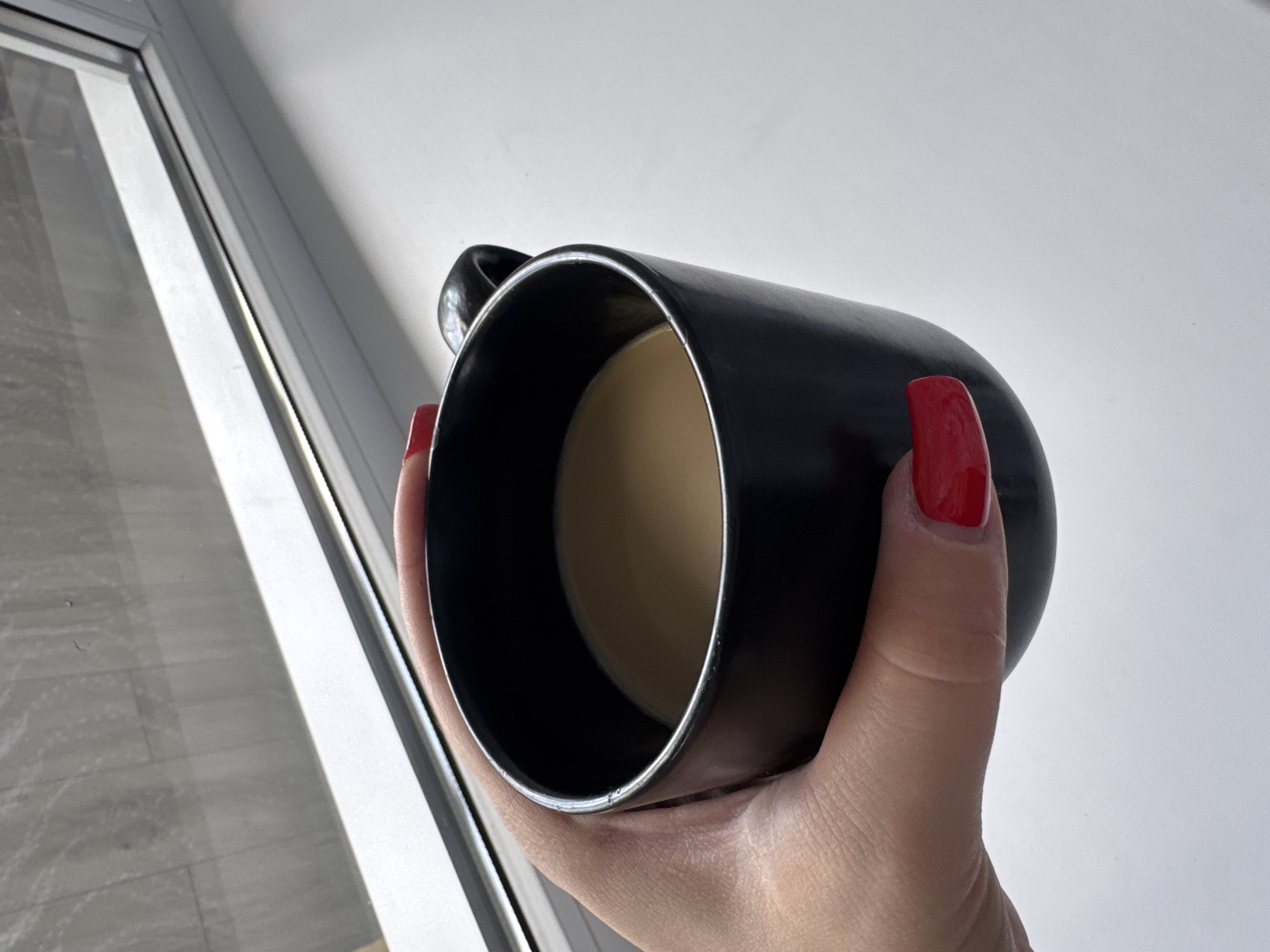 A close-up shot shows a hand with bright red nail polish holding a black mug filled with a light brown liquid, likely coffee or tea. The mug is tilted, revealing the surface of the beverage. The background is divided between a light-colored wall with diagonal shadows and a window frame with light streaming through, illuminating a floor with a grey, tiled pattern. The overall lighting suggests it might be daytime, with sunlight entering from the window. The scene evokes a sense of a quiet, personal moment, possibly during a break or while enjoying a drink by a window.