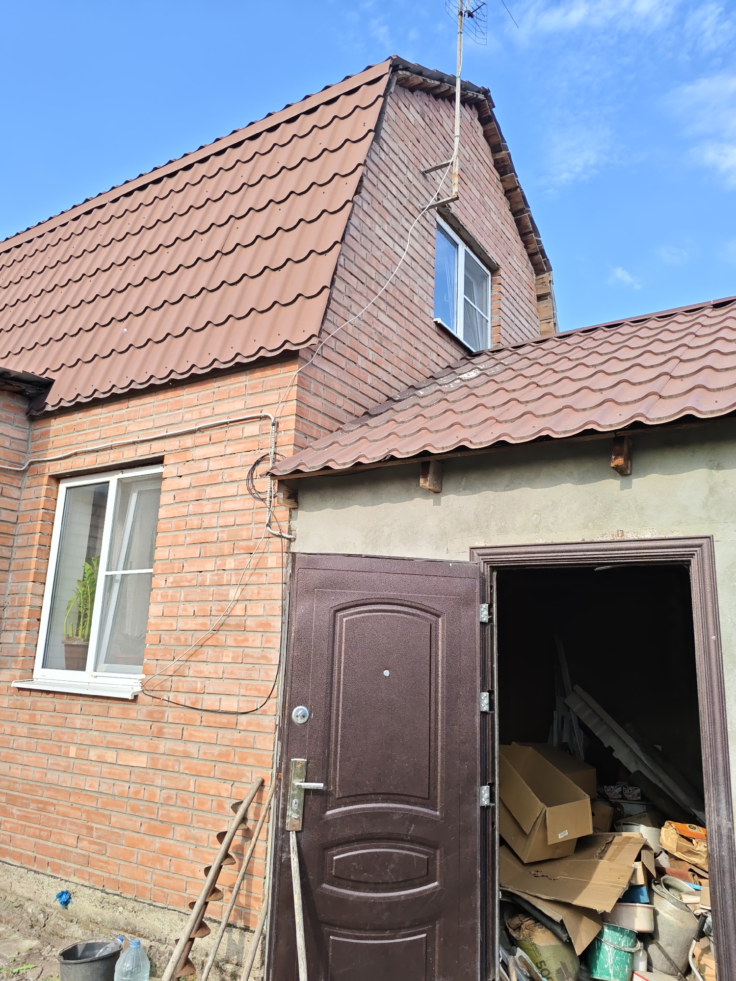 An exterior, low-angle, wide shot captures a residential brick building under a clear blue sky with a few wispy clouds in Nizhnedonskoy, Russia. The building features a brown, corrugated metal roof with a visible dormer window on the upper level. A white-framed window is present on the lower brick wall, with a potted plant visible inside. Electrical wires are run along the exterior of the building. A closed, dark brown metal door is situated to the right of the window, and next to it, an open doorway reveals a cluttered interior filled with cardboard boxes, buckets, and other assorted items. Two long, spiked tools leaning against the wall near the door.
