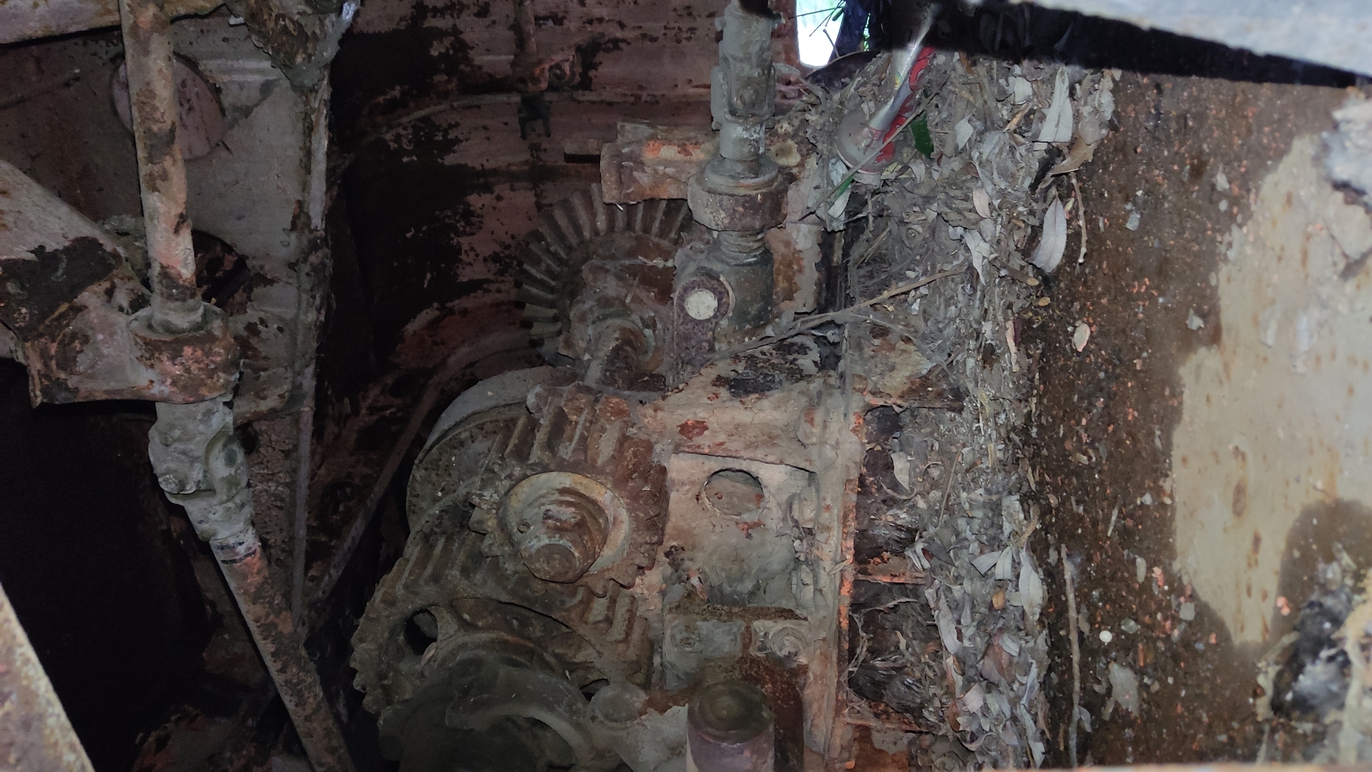 The image shows a close-up of a heavily rusted, possibly derelict, mechanical device with gears and pipes, likely an engine or machine part. It is situated in an enclosed, confined space, possibly a pit or a compartment. The surrounding environment is cluttered with debris, including leaves, twigs, and what appear to be discarded containers. The scene suggests neglect and disuse. Location information is unavailable, so no assumptions should be made.