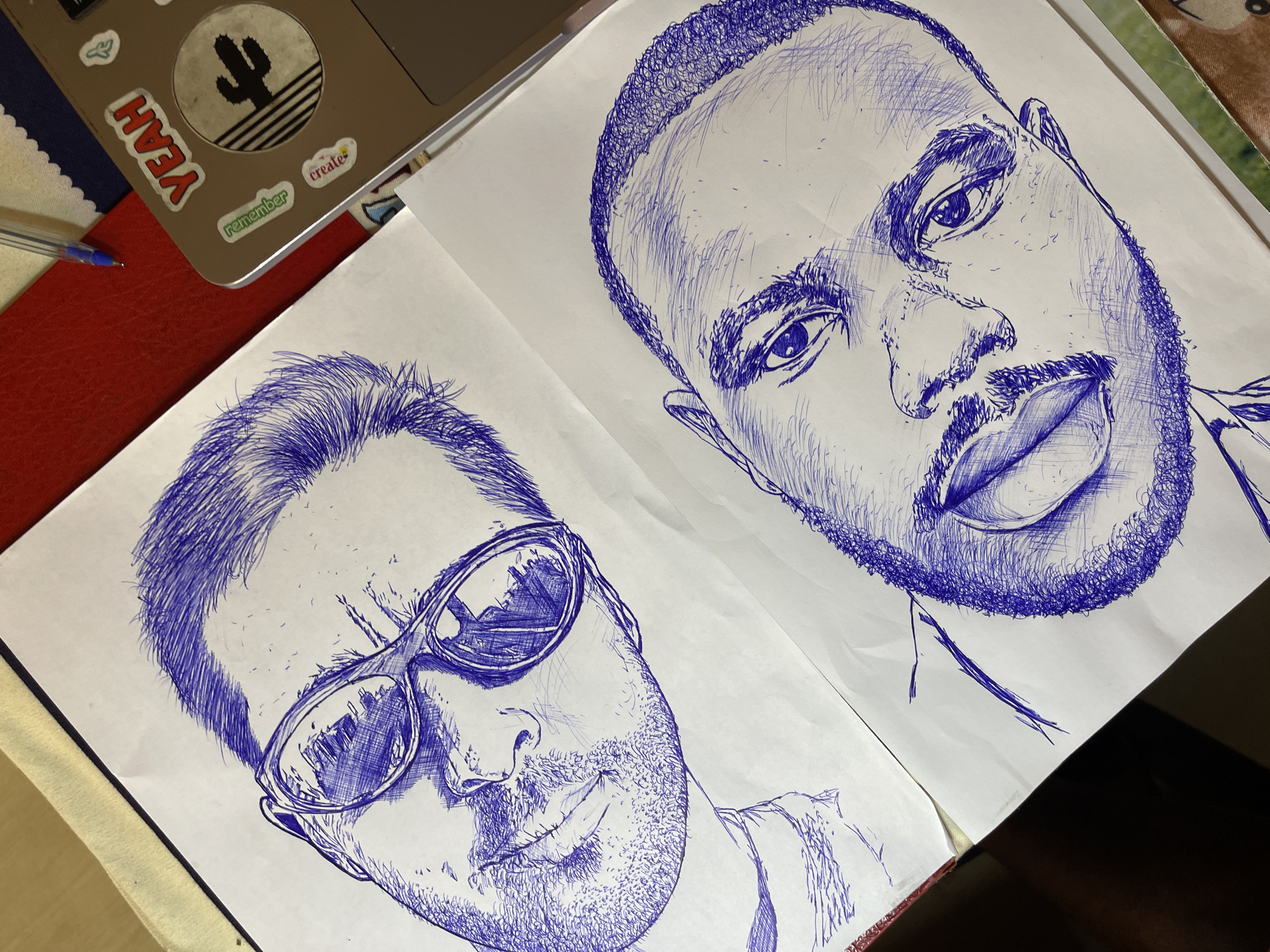 The image shows two ballpoint pen sketches of men's faces on white paper, laid out on a wooden surface. The sketch on the left depicts a man wearing sunglasses, with short hair and stubble. The sketch on the right shows a man with dark, curly hair and a mustache and beard. The paper is part of a notebook, with a red binder visible on the left edge. A laptop with stickers on it, including one that says "YEAH" and another with a cactus design, is positioned to the right of the sketches. A blue pen is also visible near the laptop. The overall scene suggests an artist's workspace. The image does not provide any information about the city of Jos, Nigeria.