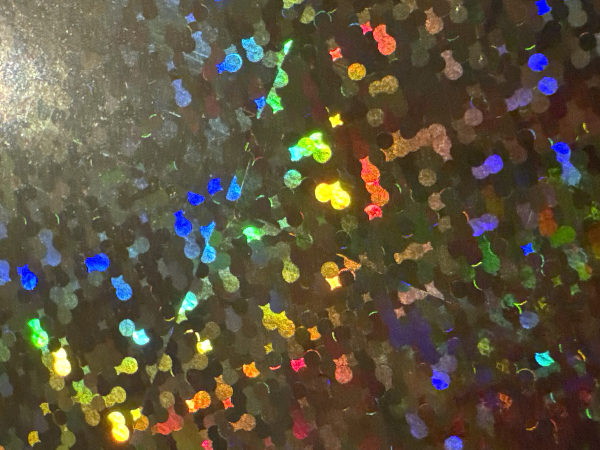 The image depicts a close-up, macro photograph of a holographic surface, characterized by a multitude of small, irregular shapes reflecting a spectrum of vibrant colors, including blues, greens, yellows, oranges, and reds against a dark, textured background. The holographic effect creates a sense of depth and shimmer, with each shape appearing to be a distinct, light-refracting element. There is no discernable connection to the city of Waterford, Ireland, as the image focuses solely on the abstract visual qualities of the material. The scene is devoid of any people, specific objects, or actions, offering a purely aesthetic and textural observation of the holographic pattern.