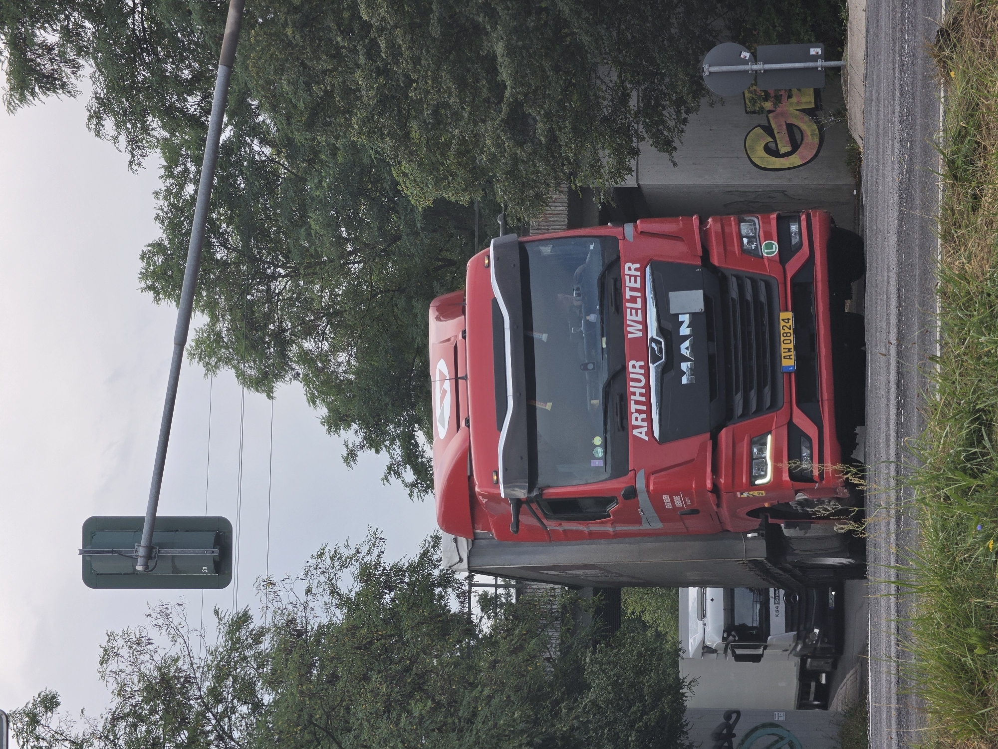 A red MAN truck with "ARTHUR WELTER" written on it is driving on a road. The truck has a gray trailer attached. In the background, a white truck is partially visible. There is a traffic light hanging from a pole over the road. Trees and a concrete bridge are also visible. Graffiti can be seen on the concrete structure. The scene appears to be in Kelsterbach, Germany, near the airport. The sky is overcast.