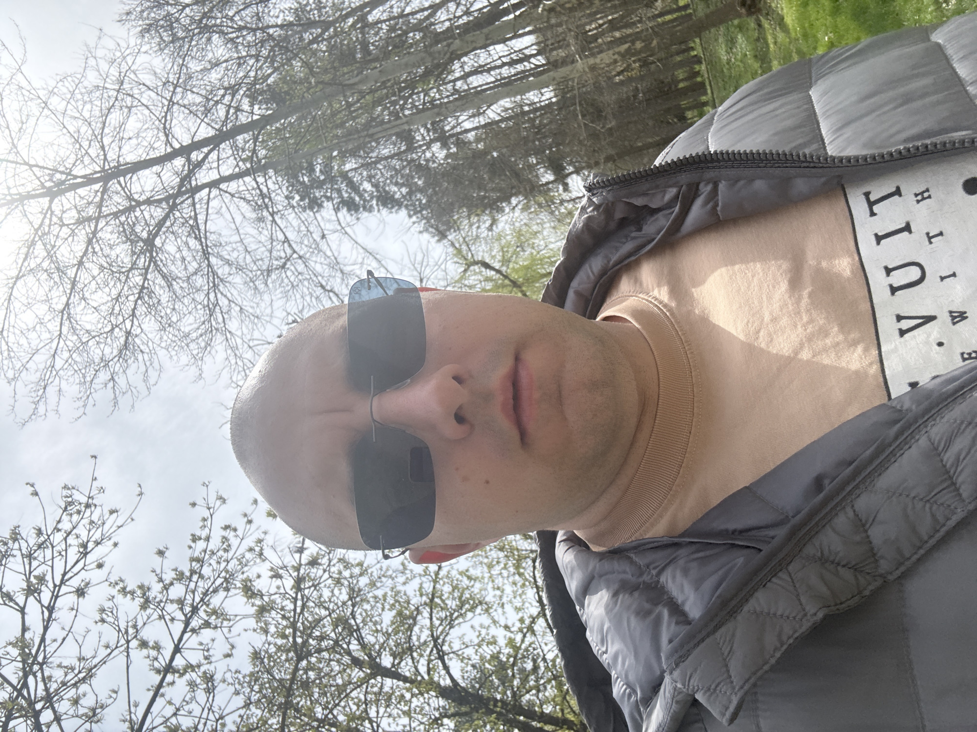A close-up, slightly angled photograph shows a bald man wearing sunglasses and a grey puffer jacket over a light brown shirt. Part of the shirt has the word "VUITT" printed on it vertically. He is looking directly at the camera. The background consists of trees with bare branches and some green vegetation, likely indicating the beginning of spring. The location is outdoors, possibly a park or wooded area. There are no readily identifiable landmarks in the image to specifically pinpoint the location within Pleven, Bulgaria.