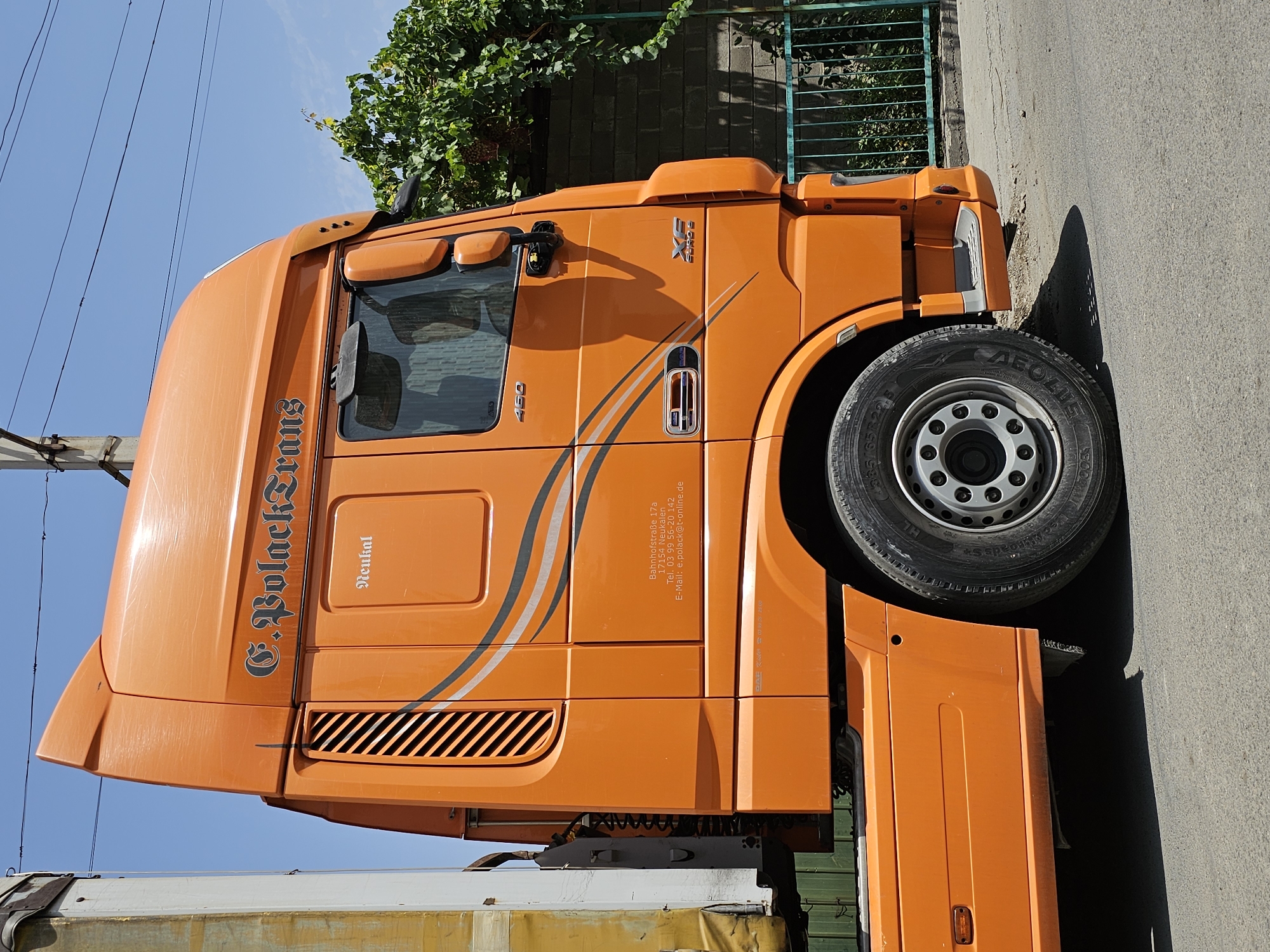 An orange DAF XF Euro 6 truck is parked on a paved road in an outdoor urban setting. The truck is viewed from the side, with its front portion closest to the camera. The truck's cab is prominent, featuring the company name "C. Bolack Trans" in stylized lettering. The number "460" is visible on the side of the cab, likely indicating engine power. A smaller label reads "Neukal". Below this, there is text indicating an address: "Bahnhofstraße 17a", "17154 Neukalen", "Tel. 03 99 56-20 142", and "E-Mail: p.polack@t-online.de". The truck has large wheels with visible tire treads and silver rims. To the left of the truck, a portion of a white and yellow trailer is visible. In the background, there is a brick wall, a green metal fence, and lush green foliage, possibly a grapevine. The sky is clear and blue. The scene suggests a sunny day. The location is specified as Özgön, Kyrgyzstan.