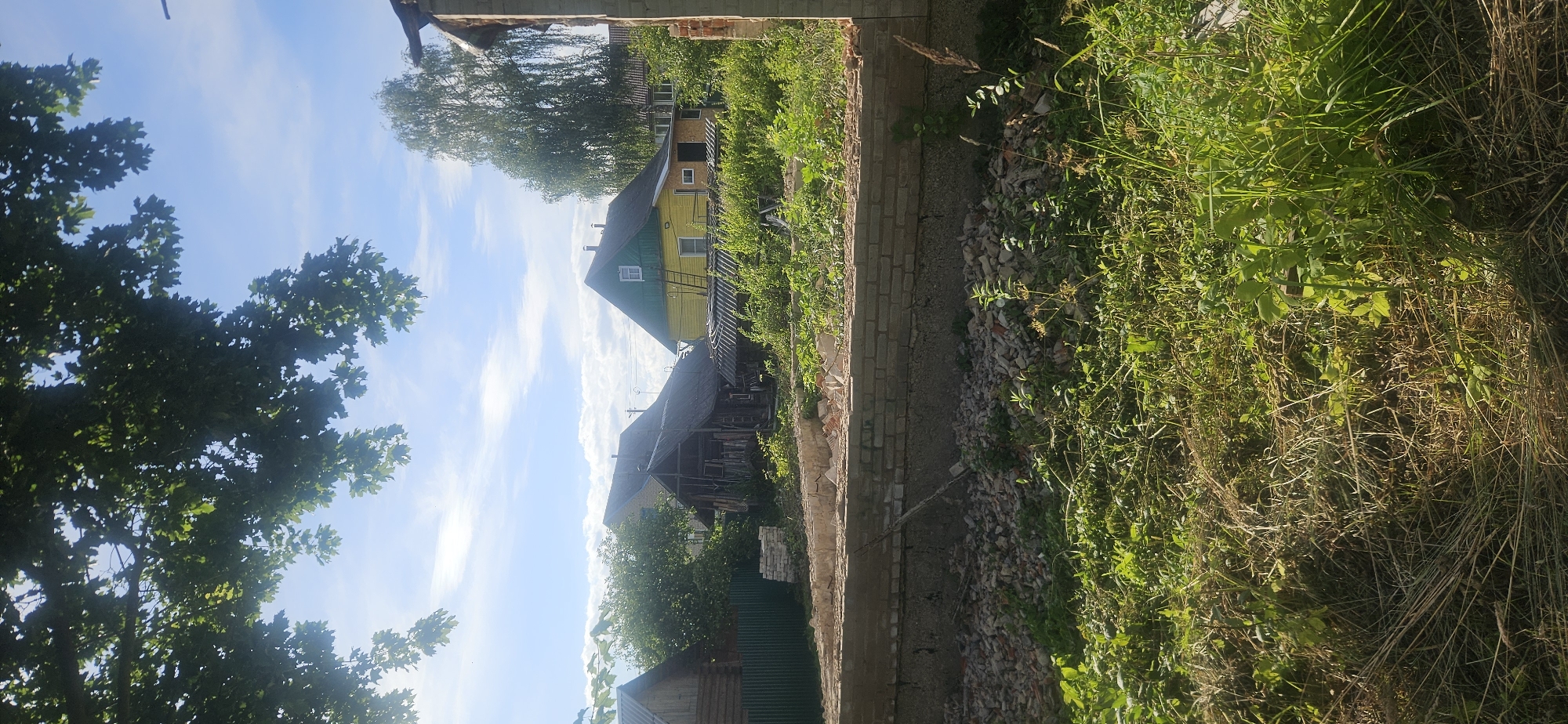 The image depicts a sunny outdoor scene in what appears to be a rural or suburban area, possibly in the city of Vitsebsk, Belarus. In the foreground, a partially constructed or demolished brick foundation is visible, with rubble and overgrown vegetation surrounding it. Several houses are situated in the background, including a yellow house with a green roof, an older, dilapidated wooden structure, and a section of a wooden fence. A large tree with green foliage is prominent in the upper left corner of the frame, with a bright blue sky and scattered white clouds above. The overall impression is one of a construction site or an abandoned property in a natural setting. There are no people or animals visible in the image, and no specific actions or interactions are depicted.