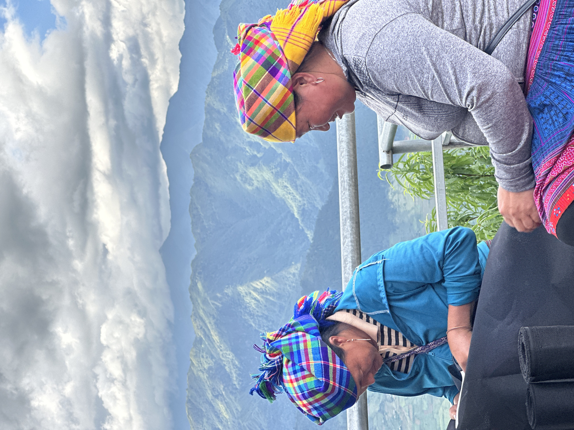 Two adult females are seated outdoors on an elevated platform with a panoramic view of mountains, likely in Tu Le, Vietnam. A grey metal railing spans horizontally between the subjects and the background.
The woman on the left wears a vibrant, multi-colored plaid headscarf predominantly featuring blue, green, and red hues, tied high with decorative tassels. She has a gold drop earring in her visible ear and a gold-colored bracelet on her right wrist. She is dressed in a light-colored striped shirt beneath a teal long-sleeved jacket. Her head is bowed as she focuses on a piece of black fabric or material laid out on a surface in front of her, holding a white tool, possibly scissors, in her right hand. Two rolls of black material are visible to her left on the same surface.
The woman on the right wears a bright, multi-colored plaid headscarf with yellow, orange, and purple as dominant colors, featuring fringes at the back. She has a silver hoop earring in her visible ear and is wearing a grey long-sleeved top with a subtle patterned texture on the shoulders. Below her waist, a traditional patterned skirt in shades of pink, blue, and red is visible. She is looking to her left with a slight smile on her face.
In the background, a vast expanse of lush, green mountains recedes into a hazy blue distance under a partly cloudy sky. Patches of white and grey clouds are interspersed with visible blue sky. Green foliage is present behind the railing between the two women.