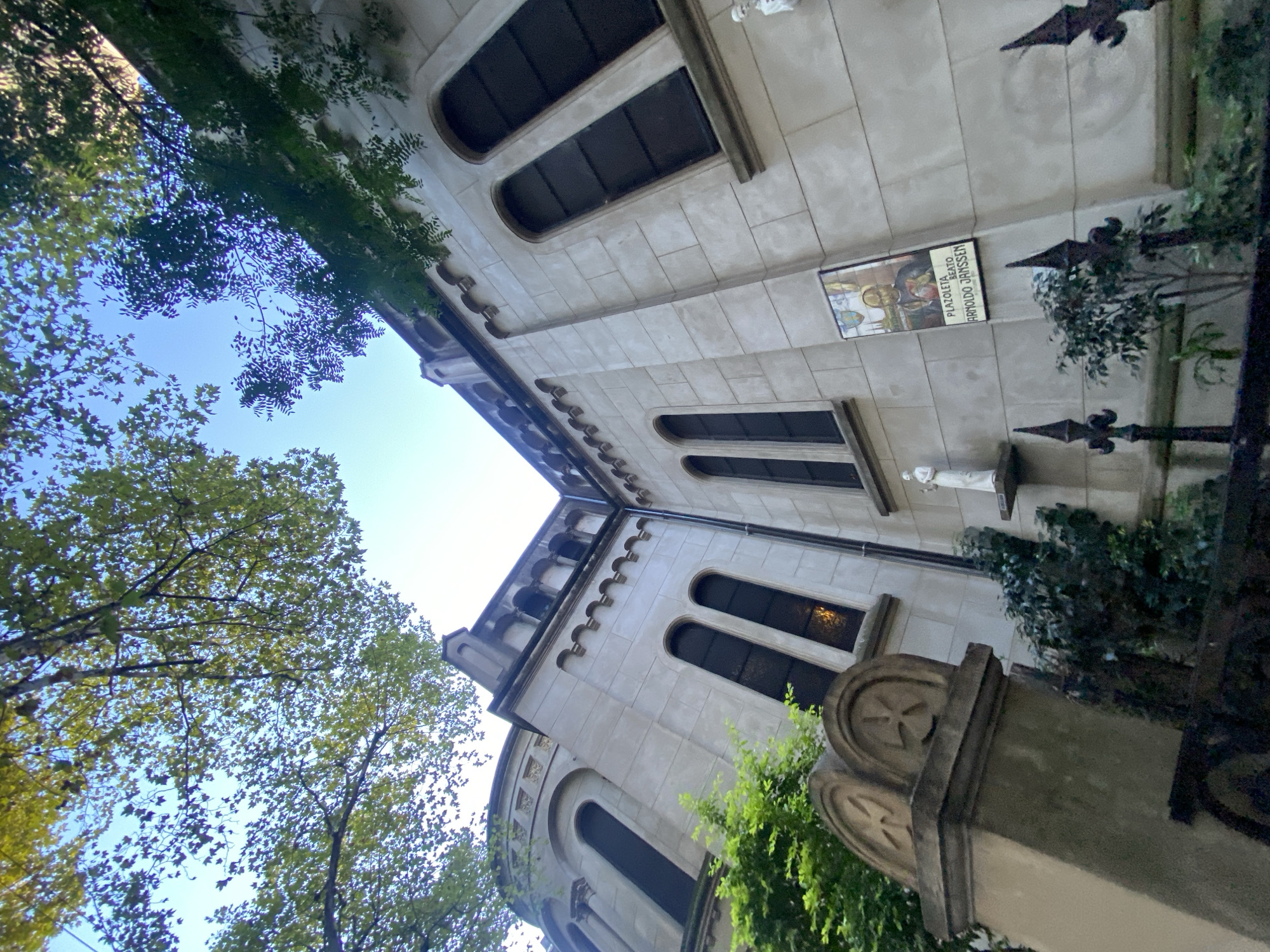 The image shows a low-angle view of the facade of a building with trees partially obscuring the view. The building is likely a church or other religious structure, judging by its architecture, including arched windows, decorative stonework, and a plaque with a religious figure. The plaque bears the words "Plazoleta Beato Arnoldo Janssen." The building is constructed of light-colored stone or concrete and appears to be in an urban environment, with trees suggesting it is located in a park or near a street lined with trees. The presence of a wrought iron fence adds to the architectural detail. The plaque refers to Arnoldo Janssen, suggesting the location is associated with a Catholic religious order or institution. Given that the language on the plaque is Spanish, the location is likely in a Spanish-speaking country. Based on the description on the plaque, the location is likely in Buenos Aires, Argentina.