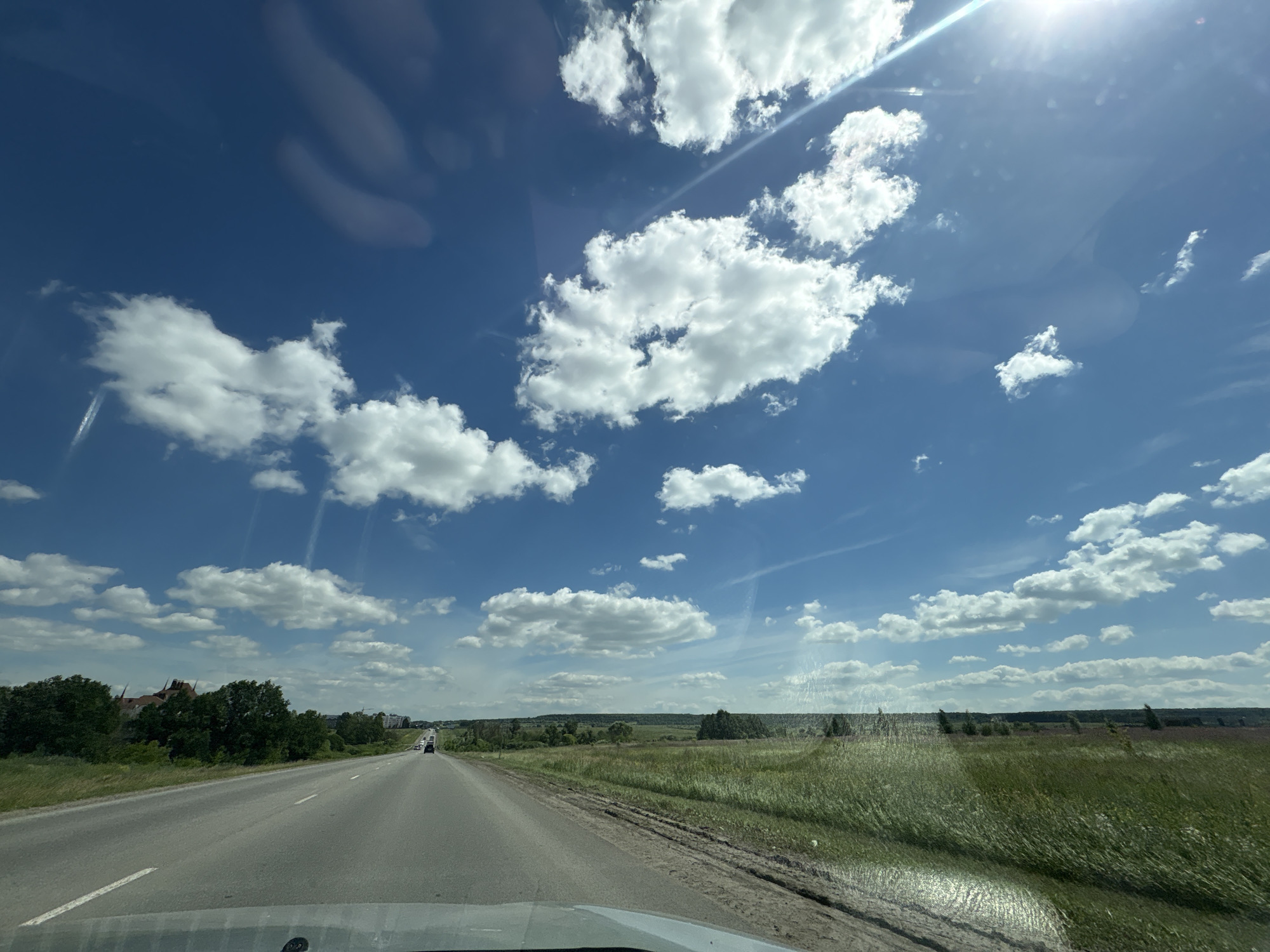 A wide-angle, slightly elevated view captures a bright, sunny day from inside a vehicle, looking forward along a paved road that curves gently to the left. The road is flanked by grassy verges and fields of green grass and some vegetation. The sky is a vibrant blue, filled with numerous fluffy white cumulus clouds. The sun is high in the sky, casting a bright glare and some lens flare. In the distance, along the left side of the road, there are trees and buildings visible, suggesting a rural or suburban setting. A few vehicles are seen on the road ahead. The overall impression is one of travel on a clear, summer day.