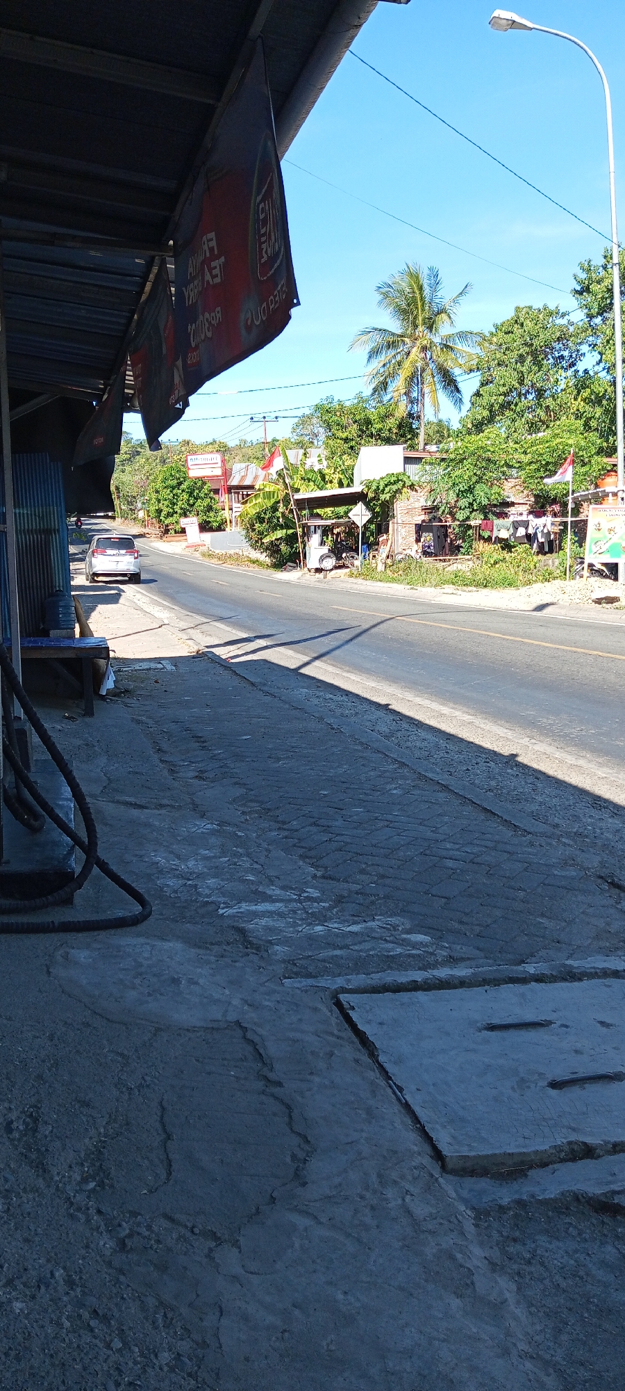 This image depicts a roadside scene in the city of Pakkanna, Indonesia, on a clear day. The foreground shows a paved walkway, likely a sidewalk, with a section of exposed concrete and a metal grate. A dark hose is coiled on the ground near the walkway's edge. Beyond the walkway is a road, with a white sedan driving away from the viewer. The right side of the road features a small roadside stall or shop with a corrugated metal roof. Various items are displayed outside the stall, including what appear to be clothing hanging to dry, a bicycle, and possibly a small water dispenser. Indonesian flags are visible, one attached to the stall and another further down the road. Lush green trees and vegetation line the opposite side of the road, with a prominent palm tree visible. In the distance, hills are visible under a bright blue sky. A street light is positioned on the right side of the image. Two red and black banners are hanging from an overhang on the left side of the image, partially obscuring the view of a blue corrugated metal structure behind them. The overall impression is of a typical rural or suburban road in Indonesia, with local businesses and homes lining the street.