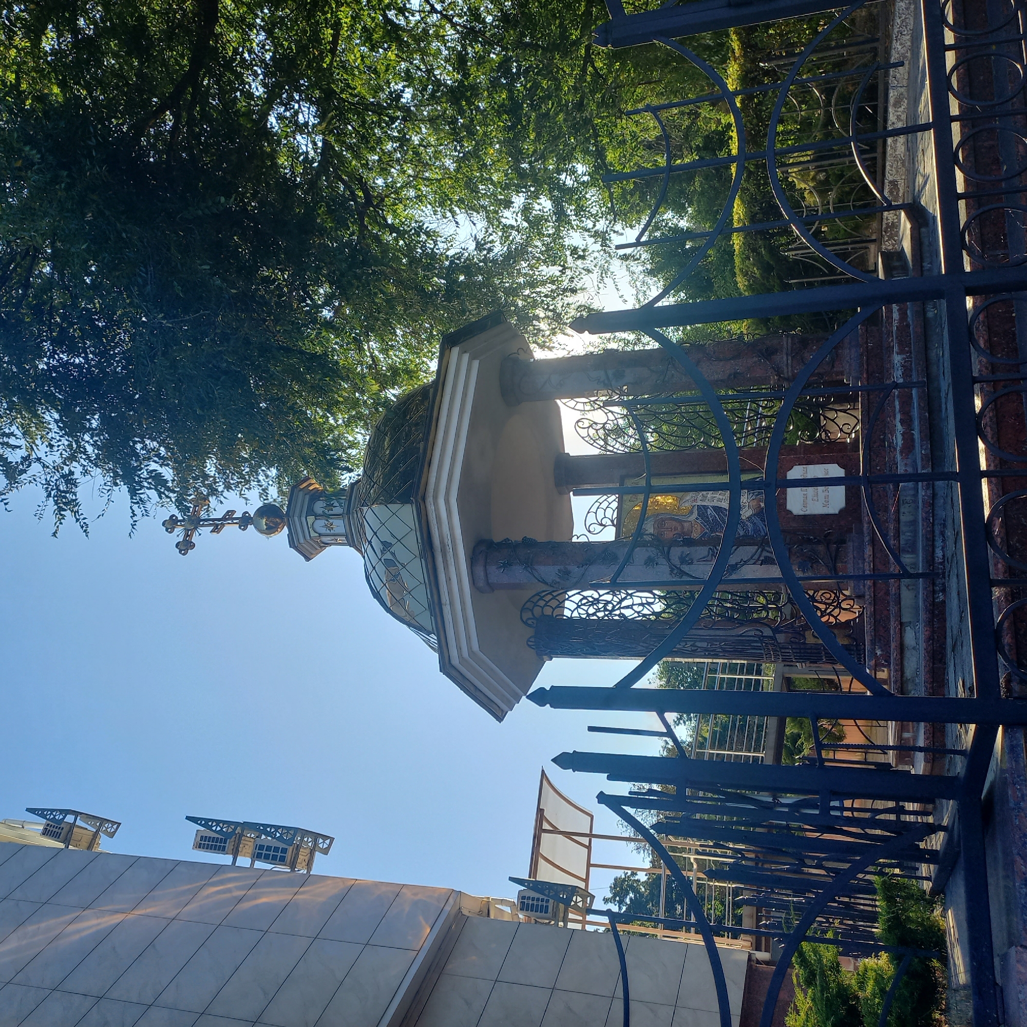 The image shows a small, octagonal structure with a dome topped by a cross, located outdoors. The structure is adorned with columns and ornate metalwork, and appears to house a mosaic of a religious figure. A metal fence encloses the structure. To the left of the structure is a modern building with air conditioning units mounted on its facade. Trees and foliage are visible in the background. The scene appears to be set during daytime with a clear blue sky. This structure is likely a chapel or shrine, possibly located within a church complex or a public space in Kropyvnytskyi, Ukraine.