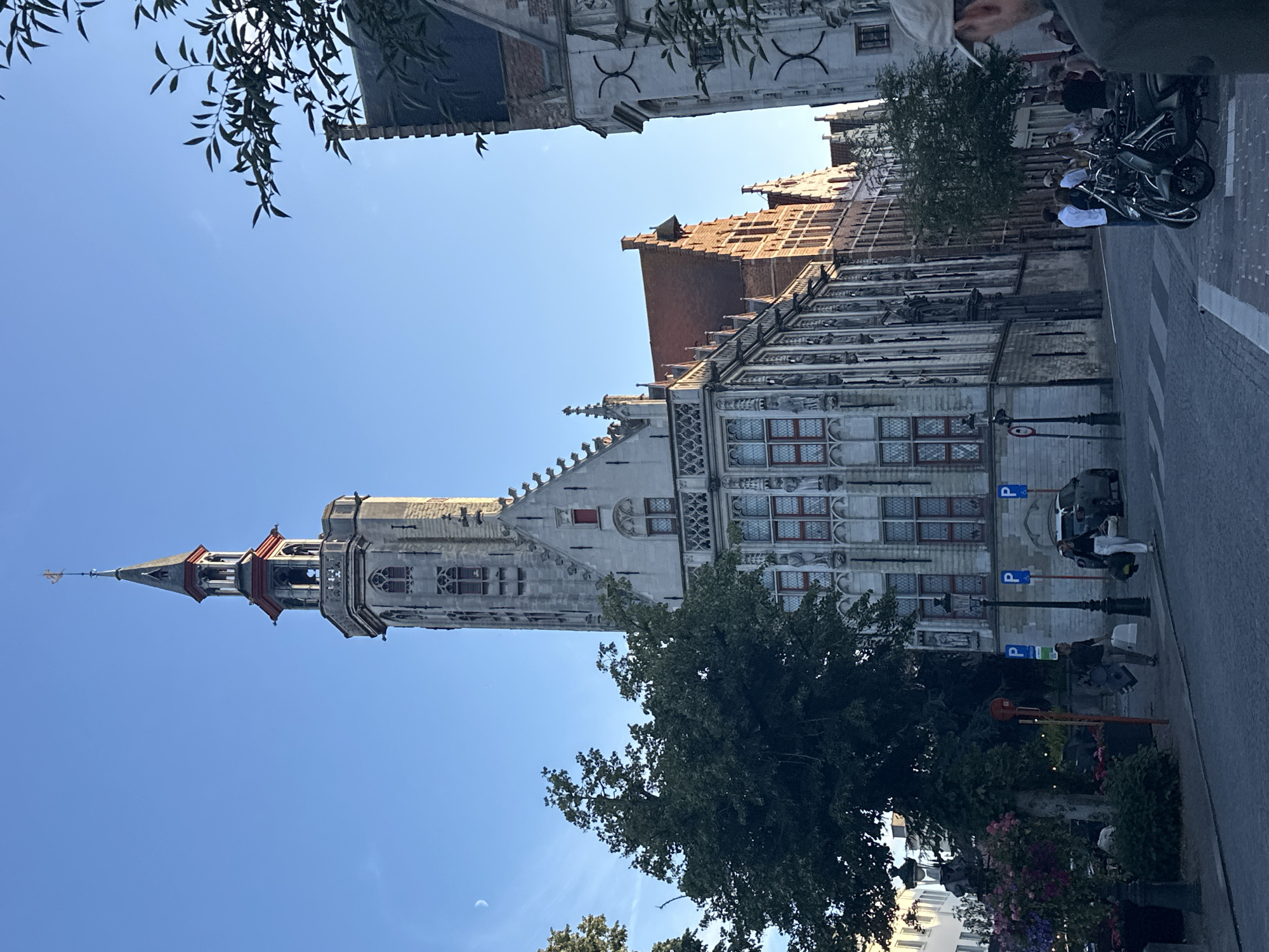 The image captures a street scene in Bruges, Belgium, dominated by historic architecture and a clear blue sky. A tall, ornate tower with a spire and a weather vane stands prominently on the left, its stone facade detailed with Gothic-style windows and decorative elements. Adjacent to the tower are several other multi-story buildings with red-tiled roofs, characterized by their stepped gables and numerous windows.
In the foreground, a street with a pedestrian crossing is visible. Several people are walking and standing on the sidewalk. Two individuals, one carrying a large, flat object and the other a dark bag, walk towards the right. A group of people are gathered near a row of parked bicycles and a scooter. Streetlights are present, and some parking signs are mounted on poles. Trees, one with lush green foliage, are interspersed along the street, and decorative flower arrangements are visible near a lamppost on the left. The scene appears to be from late afternoon or early morning, given the soft lighting and the presence of a crescent moon in the sky.