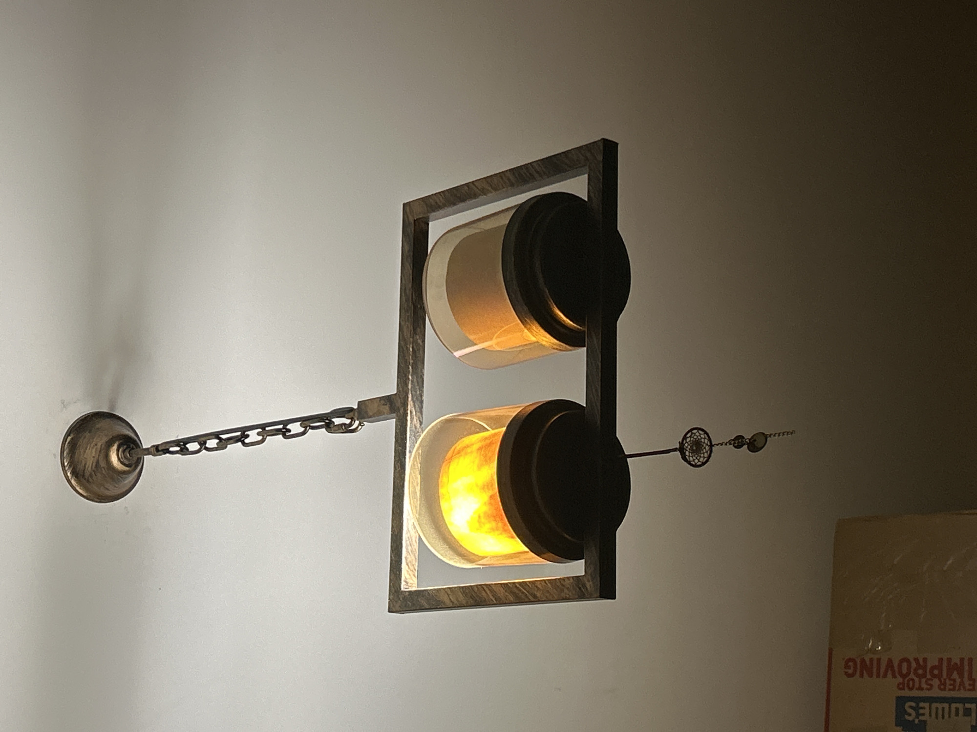An indoor shot shows a rustic chandelier hanging from a white ceiling. The chandelier has a metal frame with two cylindrical glass shades. One shade is illuminated with a warm, orange light, resembling a flame, while the other is darker and unlit. A decorative charm, resembling a small dreamcatcher, hangs from the bottom of the chandelier. In the lower left corner of the image, a portion of a cardboard box with "LOWE'S" and "IMPROVING" printed on it is visible. The overall lighting in the scene is dim, with the light from the chandelier casting shadows.