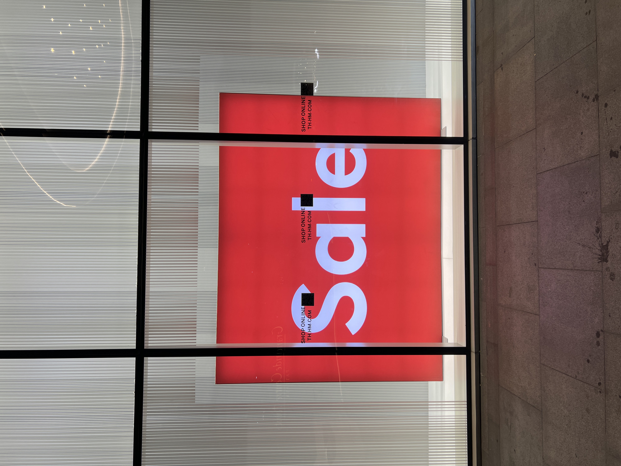 This is a nighttime exterior shot of a storefront window display. The window is divided into four panes by a black frame. Inside the window, a large red sign with white lettering announces "SALE". Below the word "SALE" are smaller white text boxes that read "SHOP ONLINE" followed by a QR code and "TH.HM.COM". The background of the display features vertical white lines. The exterior sidewalk is made of large, flat stone tiles. There are no people visible in the image.