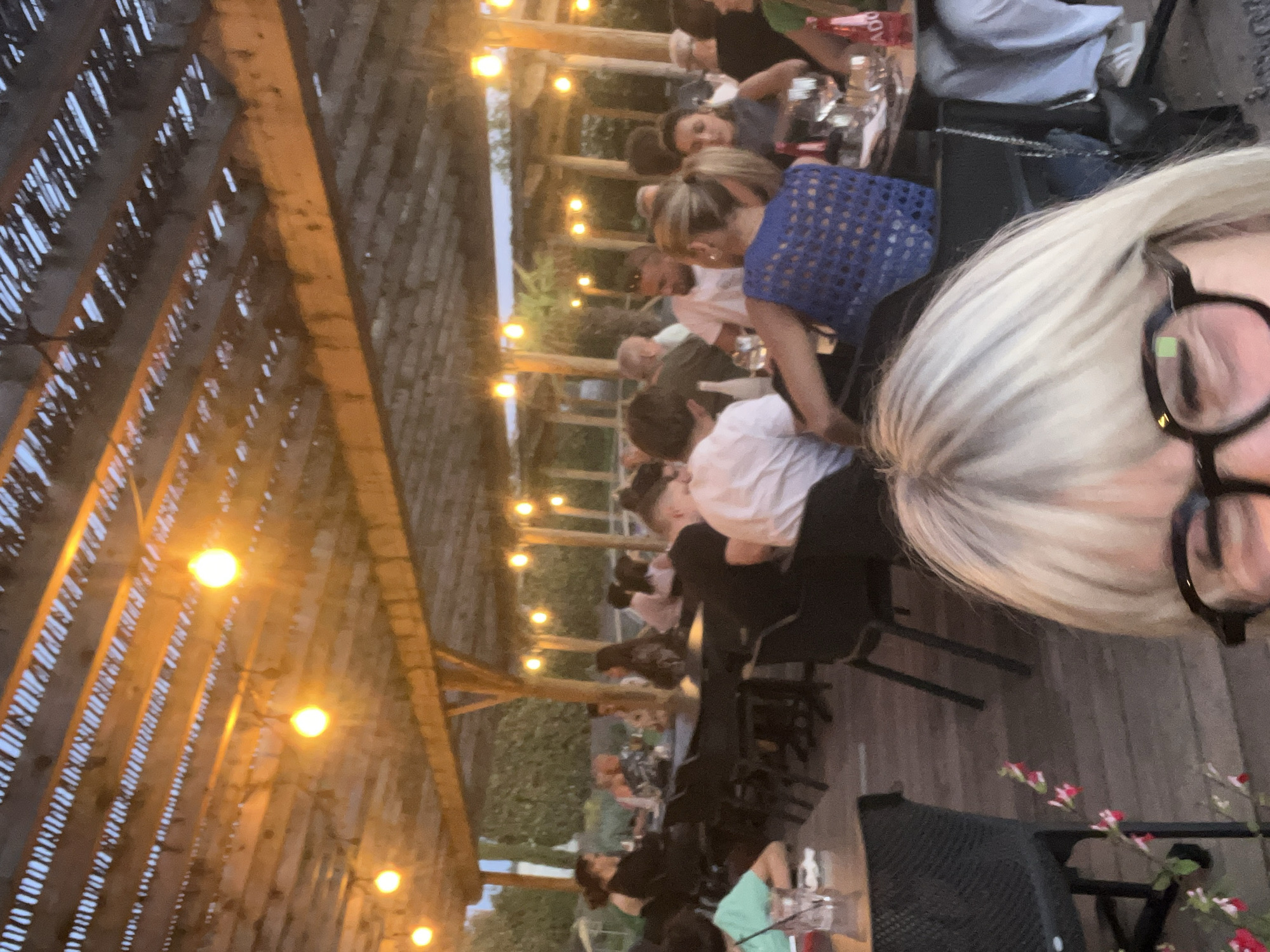 The image depicts an outdoor dining area in Andrézieux-Bouthéon, France, likely at a restaurant. Several people are seated at tables under a wooden pergola structure adorned with string lights. Diners are engaged in conversation and appear to be enjoying a meal together. The foreground features a person with blonde hair and glasses, possibly taking the photograph. Tables are set with glassware and bottles, indicating a formal dining setting. The overall atmosphere suggests a relaxed and convivial social gathering. The background reveals green foliage, suggesting an outdoor or garden setting.