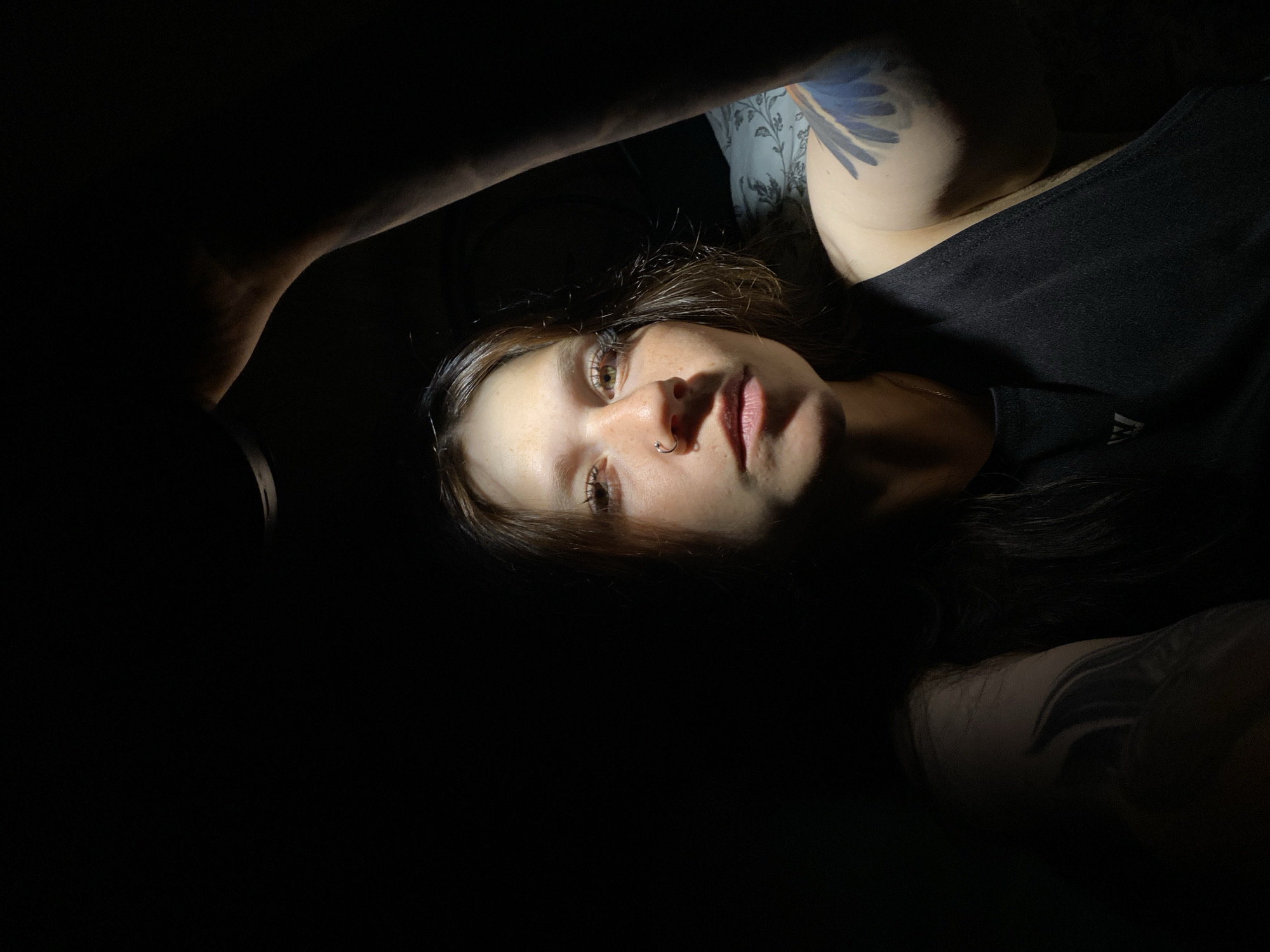 A woman is illuminated by a single light source held above her. She is looking directly at the camera with a neutral expression. Her left arm is raised, obscuring part of her face and the light source. She has a nose ring and freckles. She is wearing a black tank top. A tattoo is visible on her left arm. The background is dark and appears to be indoors, possibly a bedroom with patterned bedding visible behind her right shoulder. The scene does not provide any information about the city of Chaikovsky, Russia.
