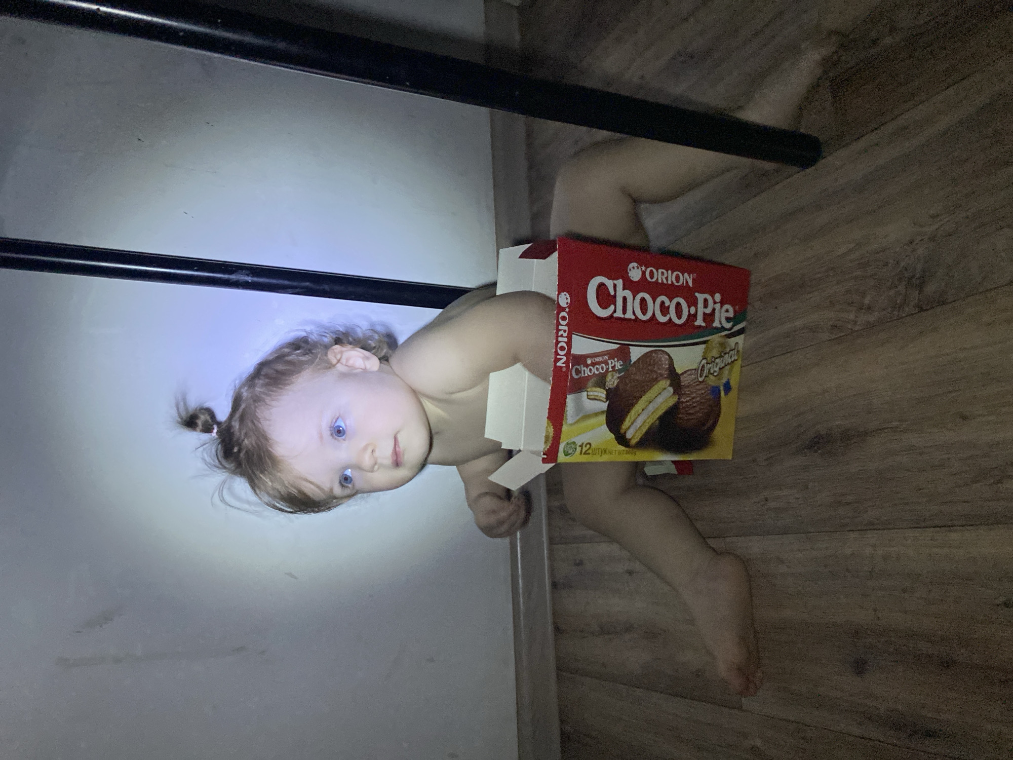 The image shows a child lying on their back with a box of Orion Choco-Pie resting on their torso. The child has fair skin, blue eyes, and light brown hair pulled into a small ponytail. The child is positioned on a boundary between a white wall and a wooden floor. Two black bars are positioned horizontally in the upper part of the frame, obscuring the view of the wall. The box is red and yellow with a picture of a Choco-Pie on it. The environment appears to be indoors. The location is not specified.