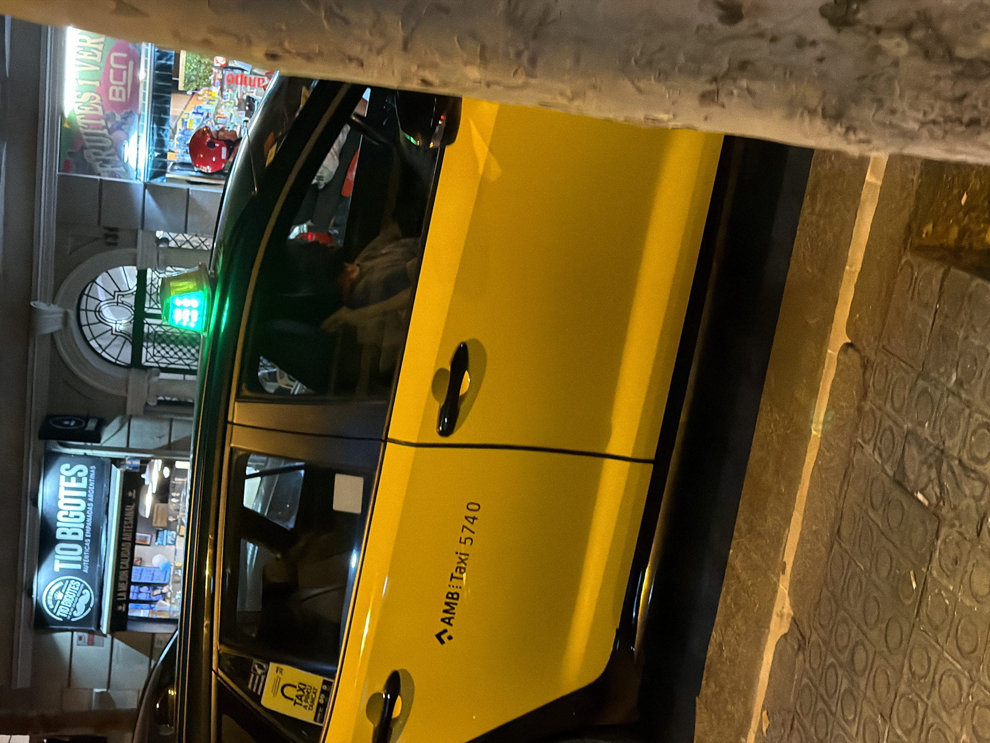 A yellow taxi with a green illuminated sign on its roof is parked on a cobblestone street at night in Barcelona, Spain. The taxi has black trim and door handles, and the text "AMB:Taxi 5740" is visible on its side. A yellow sticker with a black taxi symbol and Catalan text is affixed to the driver's side rear window. Inside the taxi, a person wearing a grey hoodie is visible in the driver's seat. To the right of the taxi, a tree trunk is partially visible, and behind the taxi, two storefronts are illuminated. One storefront displays a sign that reads "TIO BIGOTES" with text underneath indicating "AUTÉNTICAS EMPANADAS ARGENTINAS." The other storefront has a sign that says "FRUITES I VERD." with "BCN" in smaller letters. A person wearing a red helmet is visible in the doorway of the storefront to the right. The street is lit by artificial lighting, casting shadows and highlights on the scene.