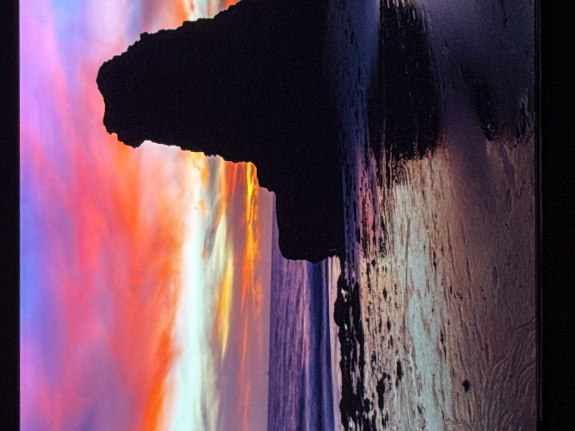 The image displays a dramatic sunset over a beach and the sea. The sky is ablaze with vibrant colors, transitioning from deep purple at the top to fiery orange and pink near the horizon, with streaks of lighter blue and white clouds. In the foreground, a dark, silhouetted rock formation dominates the right side of the frame, its jagged edges sharply defined against the colorful sky. The wet sand on the beach reflects the colors of the sunset, creating a shimmering, almost mirror-like surface. Small rocks are scattered along the shoreline. The sea is a deep, textured purple, with gentle waves lapping at the shore. The overall scene is one of natural beauty and dramatic intensity. There are no people or buildings visible, and the image appears to be a natural landscape photograph.