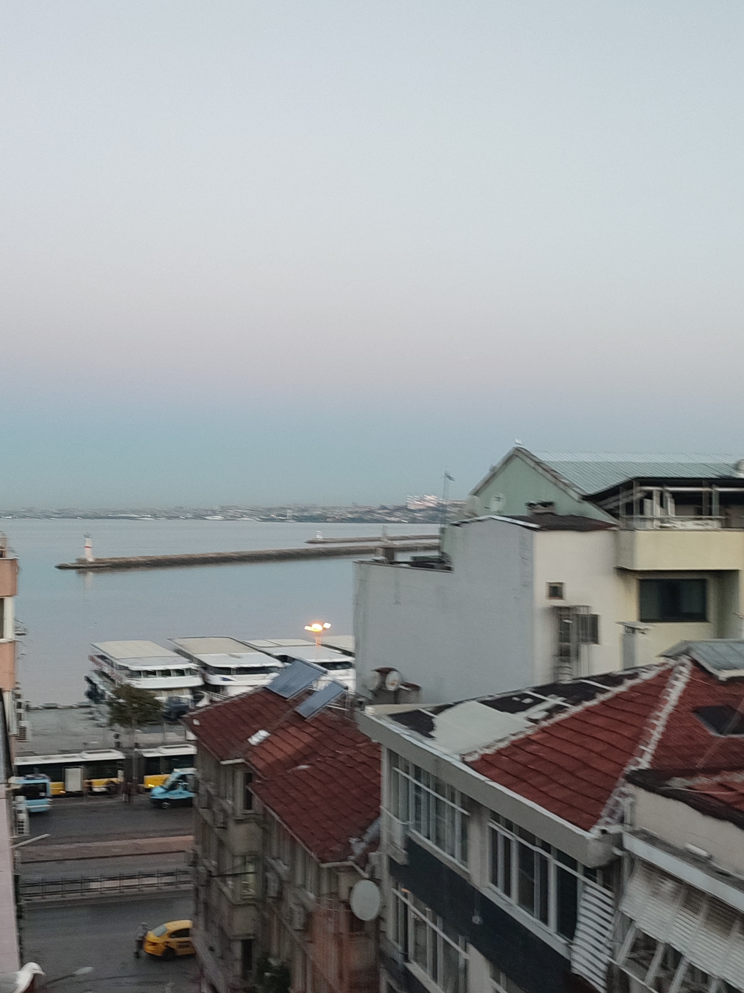 This image captures a view overlooking a waterfront area in Kadıköy, Turkey, likely from an elevated position such as a balcony or window. The scene is dominated by a body of water, possibly the Sea of Marmara, with a long breakwater extending into it, marked by a lighthouse at its end. Another lighthouse is visible further in the distance along the coastline.
In the foreground, several buildings are visible. The closest building in the lower right has a red-tiled roof and a multi-story facade with numerous windows. To its left, other buildings with varying roof types and architectural styles are present. In the middle ground, several large, white boats or ferries are docked at a pier. Across the street from the pier, there are at least two buses, one blue and one yellow, and a smaller blue vehicle. A yellow taxi is also parked on the street, with a person standing nearby.
The sky above is overcast and pale, suggesting either early morning or late evening light. The overall atmosphere is calm and urban, with a blend of maritime elements and city infrastructure. The presence of ferries and buses indicates a transportation hub, characteristic of a bustling port city like Kadıköy.
