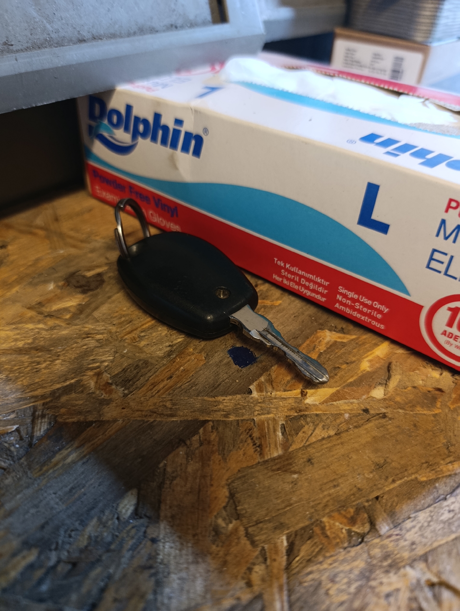 The image shows a close-up of a car key and a box of Dolphin brand vinyl gloves on a wooden surface. The wooden surface appears to be a workbench or table made of particleboard. The box of gloves is primarily white with blue and red accents, and it is marked with the brand name "Dolphin" and the size "L". Text on the box indicates the gloves are "Powder Free Vinyl," "Single Use Only," "Non-Sterile," and "Ambidextrous." The car key is black with a silver metal head and appears to be a remote key fob. The background is out of focus, but suggests an indoor environment with shelves and other objects. The scene is set in Ümraniye, Turkey.