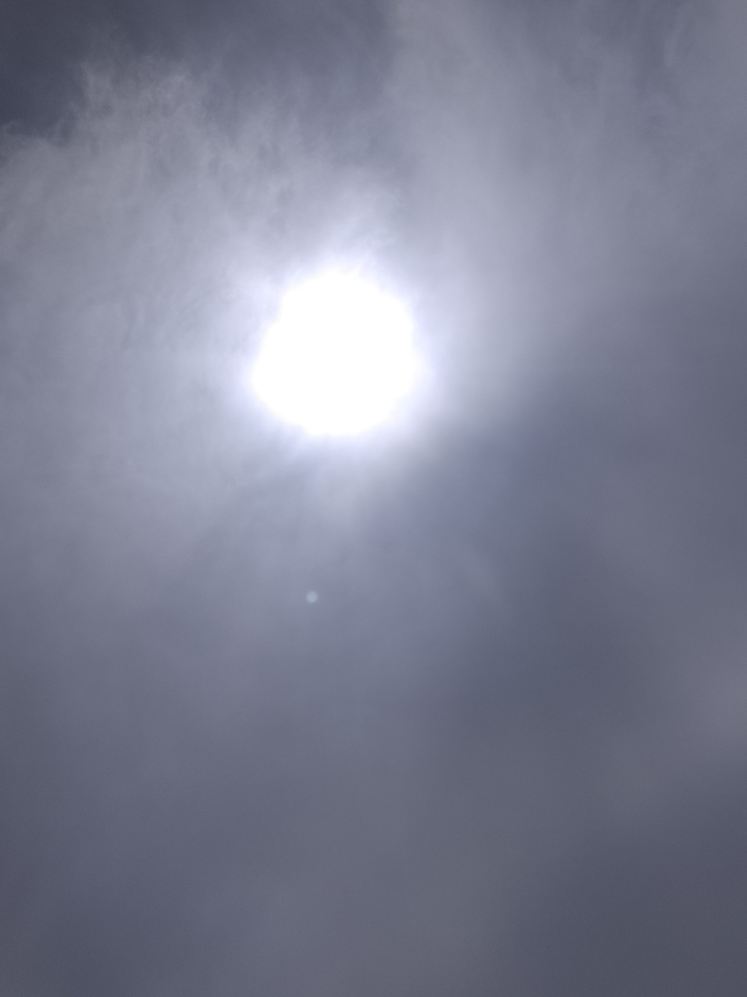 The image shows the sun obscured by clouds in Altagracia, Venezuela. The sun appears as a bright, circular area surrounded by lighter clouds, contrasting with the darker, overcast sky. A small, bright reflection or point of light is visible below the sun. The overall environment is cloudy and overcast.