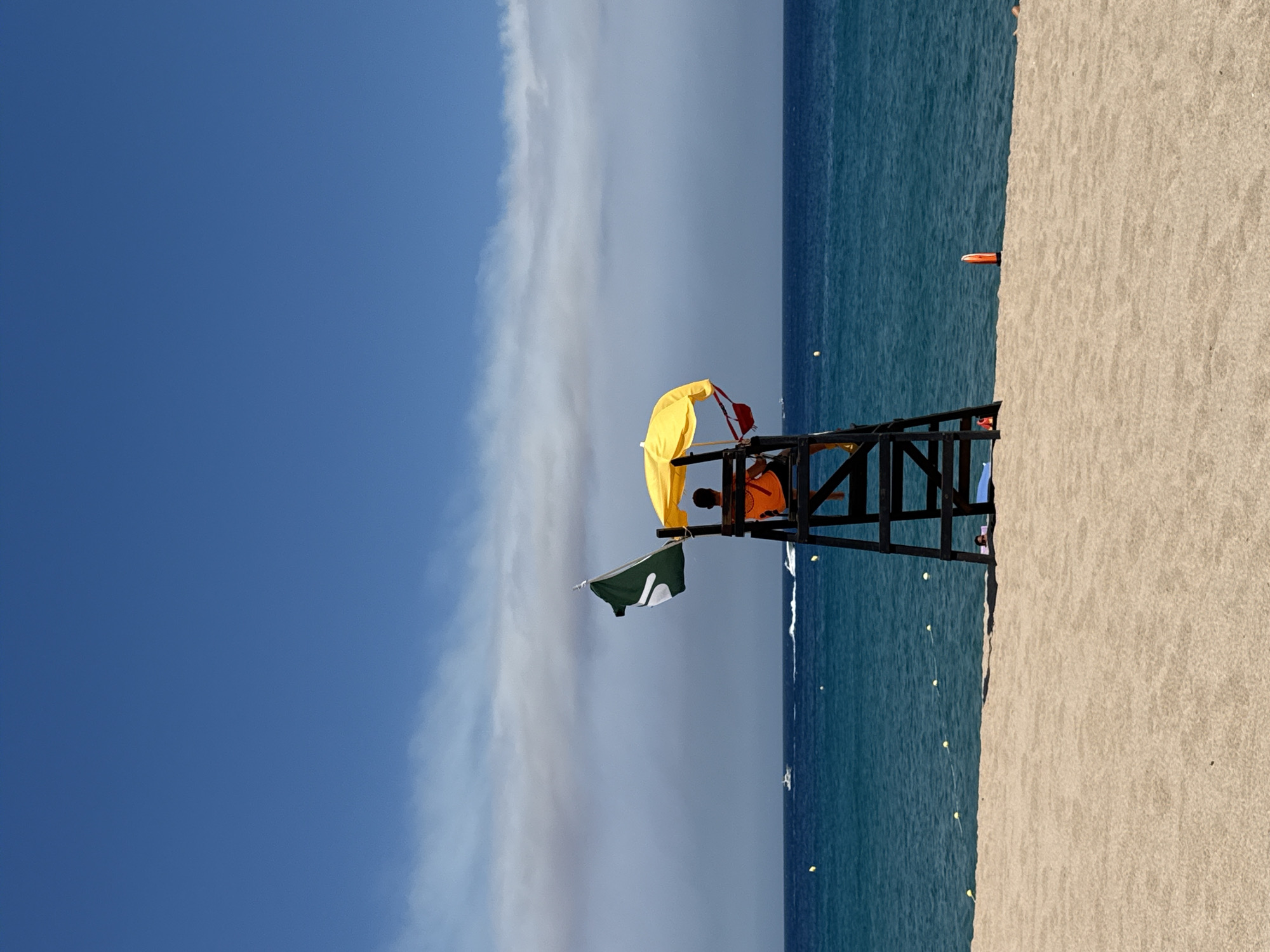The image depicts a beach scene in Blanes, Spain, on a clear, sunny day. The foreground is dominated by a wide expanse of light brown sand. In the middle ground, the turquoise blue water of the Mediterranean Sea meets the sand. A lifeguard tower, made of dark wood, stands on the beach, overlooking the water. A lifeguard, wearing an orange shirt, is seated in the tower, beneath a bright yellow umbrella. A green flag with a white symbol is attached to the lifeguard tower, fluttering in the breeze. Several yellow buoys are visible in the water, marking a swimming area. In the background, a few boats are sailing on the sea, and a hazy line of clouds is present in the sky. The overall atmosphere is one of a calm and pleasant beach day.