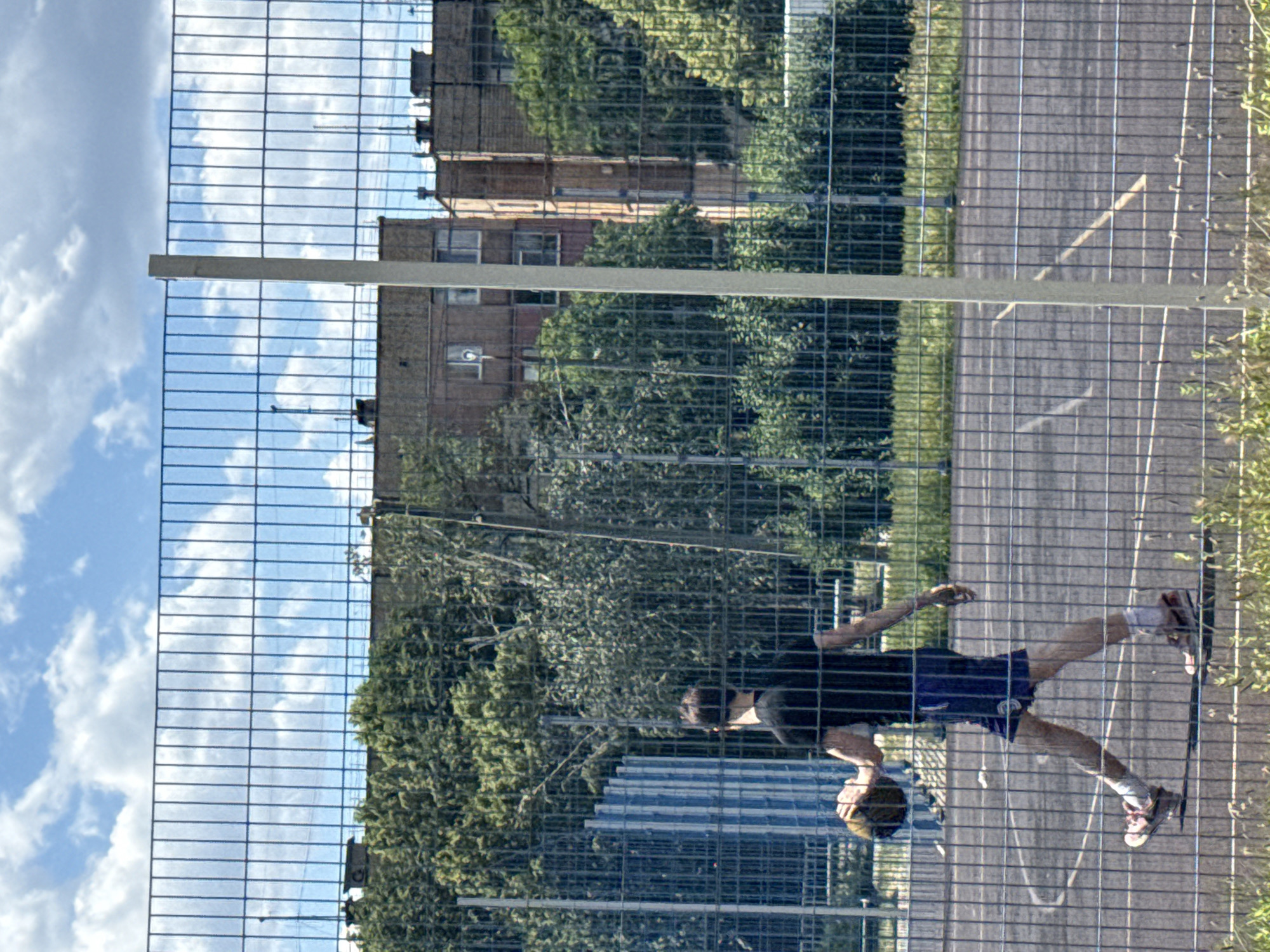 A young man wearing a black t-shirt and blue shorts walks across a basketball court, holding a brown basketball in his left hand. He is wearing white socks and pink and white sneakers. The court is fenced by a metal mesh fence, and in the background, there are apartment buildings and lush green trees under a blue sky with scattered clouds. The image appears to be taken from outside the fence, looking in.