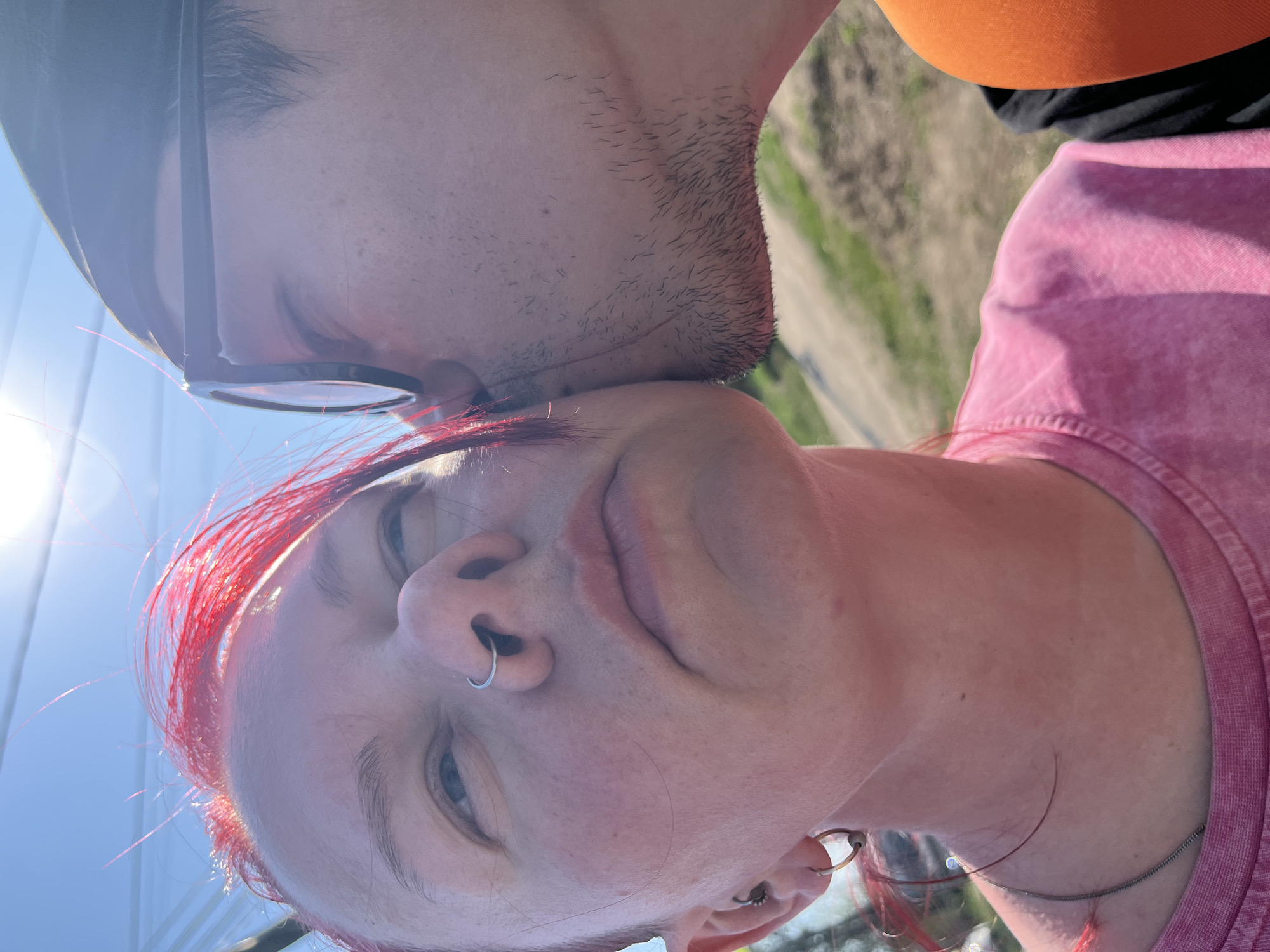 A close-up, canted, slightly low-angle shot depicts a fair-skinned person with red hair being kissed on the forehead by another individual. The person being kissed has a nose ring and multiple earrings in their left ear. The kissing individual has facial hair, is wearing glasses, and part of an orange shirt is visible. A glimpse of a landscape with a road and vegetation suggests an outdoor location. There is no definitive evidence in the image to confirm the location is Chaikovsky, Russia.