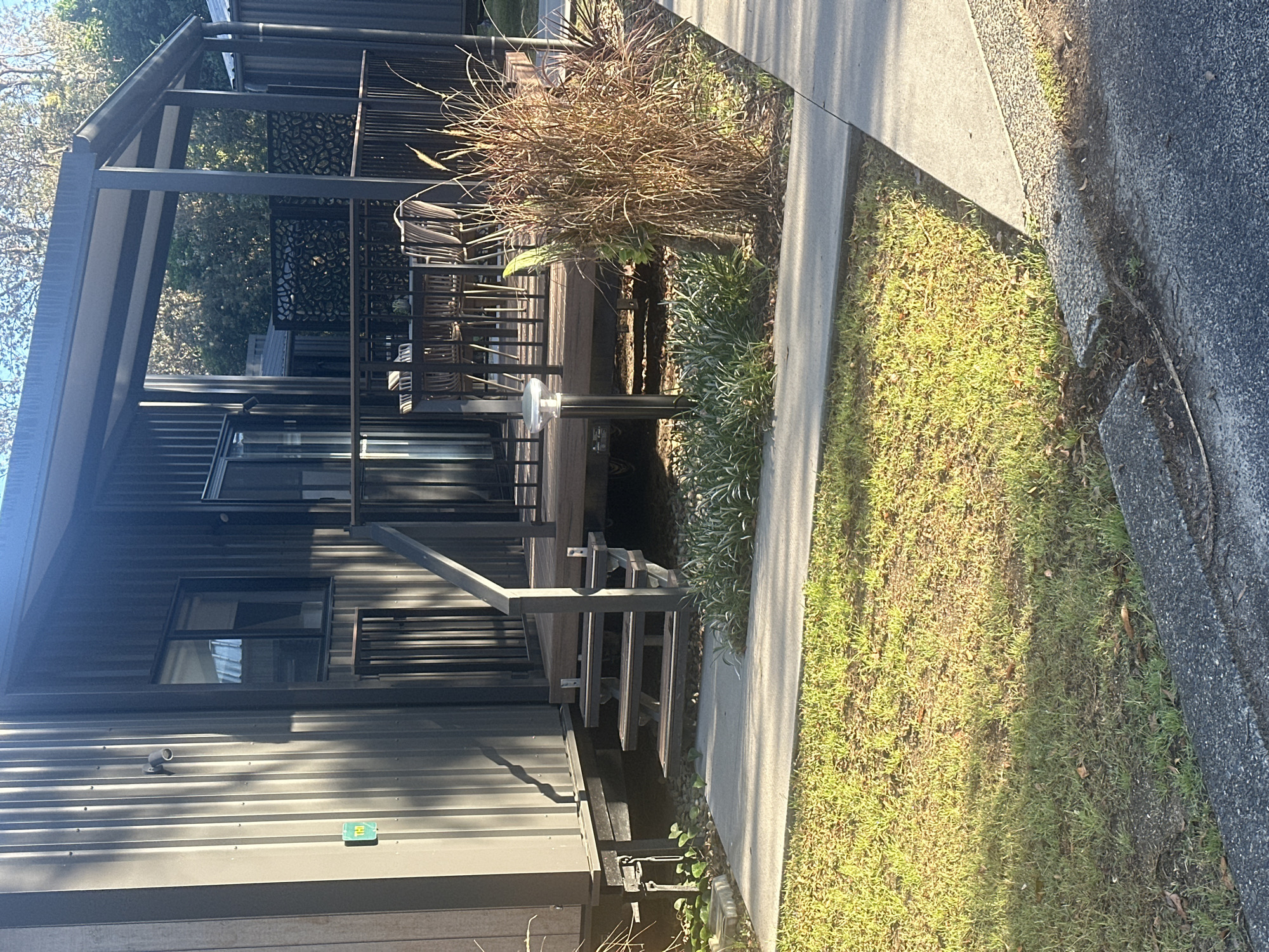 The image shows a close-up of a modern, dark gray corrugated metal tiny house or cabin with a covered porch. The cabin is situated in Mylestom, Australia. A wooden deck with black metal railings extends from the front of the cabin, accessible by a set of wooden stairs with metal treads. On the deck, a table with several chairs is visible. In front of the cabin, a paved walkway runs alongside a grassy area. Tall, dry ornamental grasses are planted to the right of the walkway, near the porch. A modern-style outdoor light fixture stands on a pole in front of the deck. The scene is illuminated by natural sunlight, casting shadows across the grass and the front of the cabin. The overall impression is that of a residential outdoor setting.