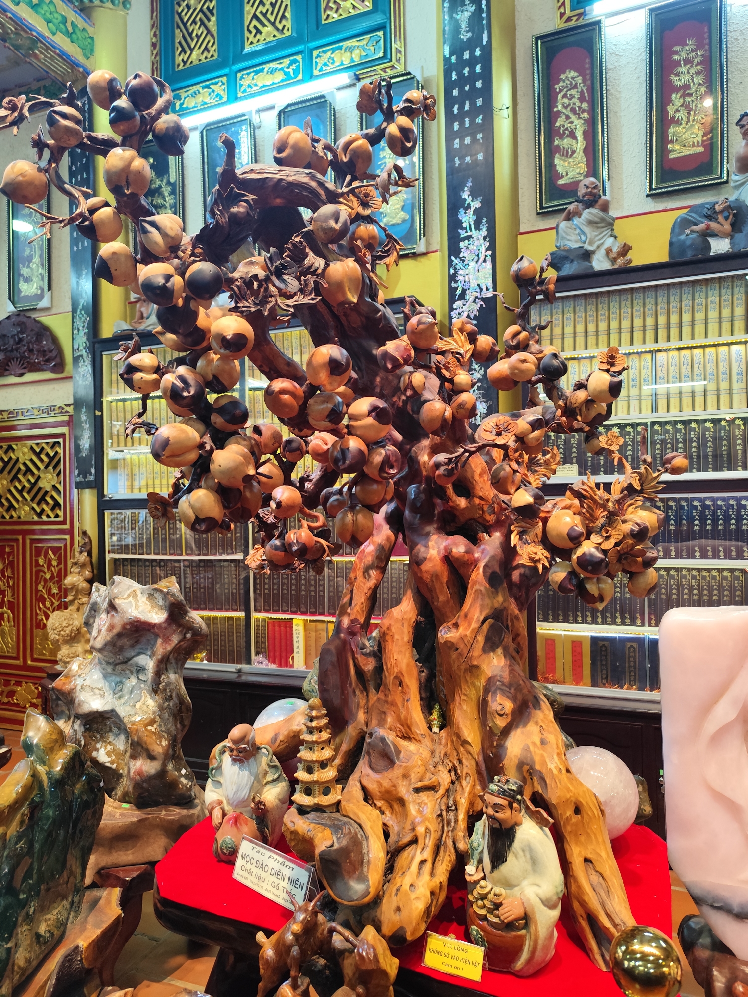 This is a photograph of a display of carved wooden sculptures and other decorative items. The centerpiece is a large, intricately carved wooden tree with numerous fruit-like ornaments. To the left of the tree, there is a textured, rock-like sculpture. In front of the tree, on a red velvet surface, are two small ceramic figurines, one depicting an elderly man with a beard holding a bag and a small pagoda, and another depicting a man holding coins and a green object. A sign on the red surface reads "Tác phẩm Mộc Đảo Diên Niên" and another sign below it has smaller text, potentially detailing the artwork. Behind the display, shelves are filled with books, and ornate framed artwork adorns the walls. The overall environment appears to be an indoor display area, possibly a shop or a gallery, with a warm, richly colored aesthetic. The location is identified as Xuân Thành, Vietnam.