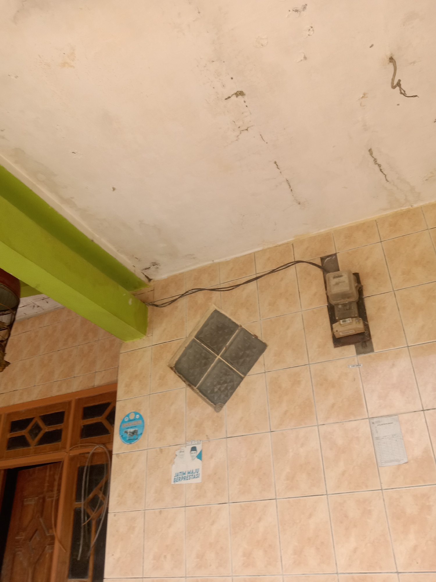 The image shows a section of a building's exterior wall and ceiling, tiled with light brown square tiles. A green beam is visible in the upper left corner. Mounted on the wall are an electrical meter box and two stickers, one circular and blue with an image, and another rectangular with text and a picture of two men. A wooden door with glass panels is seen to the left of the wall. The ceiling appears to be made of a rough, off-white material with some water stains and dark marks. The scene is likely located in Stiaji, Indonesia, given the context provided.