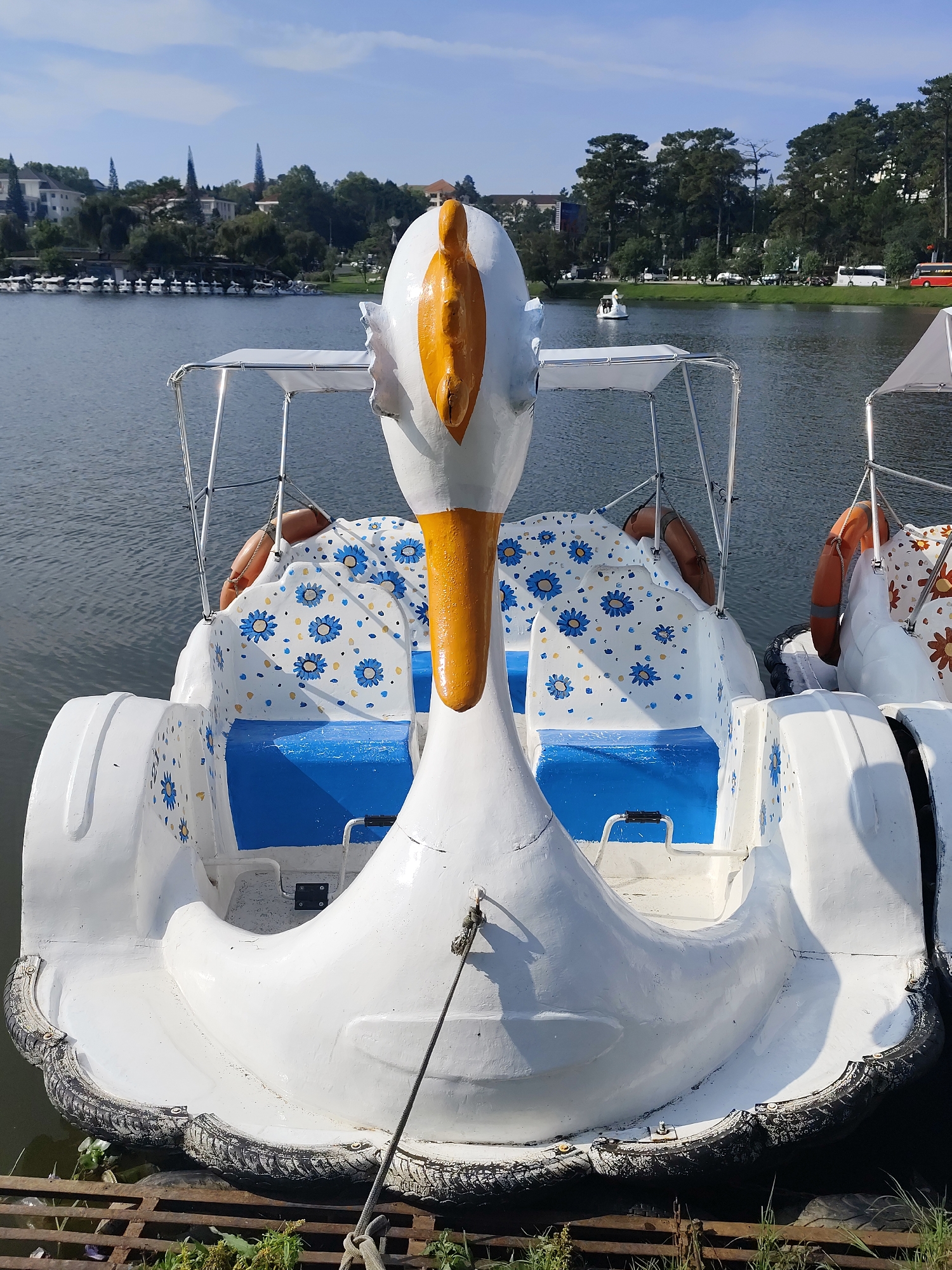 The image captures a white, swan-shaped pedal boat docked at the edge of a serene lake in Da Lat, Vietnam, on a bright, sunny day. The boat, serving as the central subject, features a prominent yellow head and beak. Its interior is adorned with white seats upholstered in a playful blue floral pattern, complemented by solid blue seat bases. Two orange lifebuoys hang from the sides, and a light-colored canopy frame provides shade over the seating area. The boat's lower perimeter is protected by a bumper made of dark, worn tires, and it is secured to a wooden and metal dock structure with a rope.
In the background, the calm waters of the lake reflect the clear blue sky with sparse clouds. Across the lake, a lush green shoreline is visible, densely covered with trees, including several distinctive tall, pointed evergreens. Behind the trees, buildings resembling houses or hotels dot the landscape. Further to the right, a road runs parallel to the lake, where vehicles including cars and buses can be seen. Other similar swan pedal boats are visible docked to the right of the main subject, and a few are scattered across the lake, indicating a popular tourist activity. The scene suggests a pleasant, leisurely atmosphere typical of a lakeside resort town. No discernible text is visible in the image.
