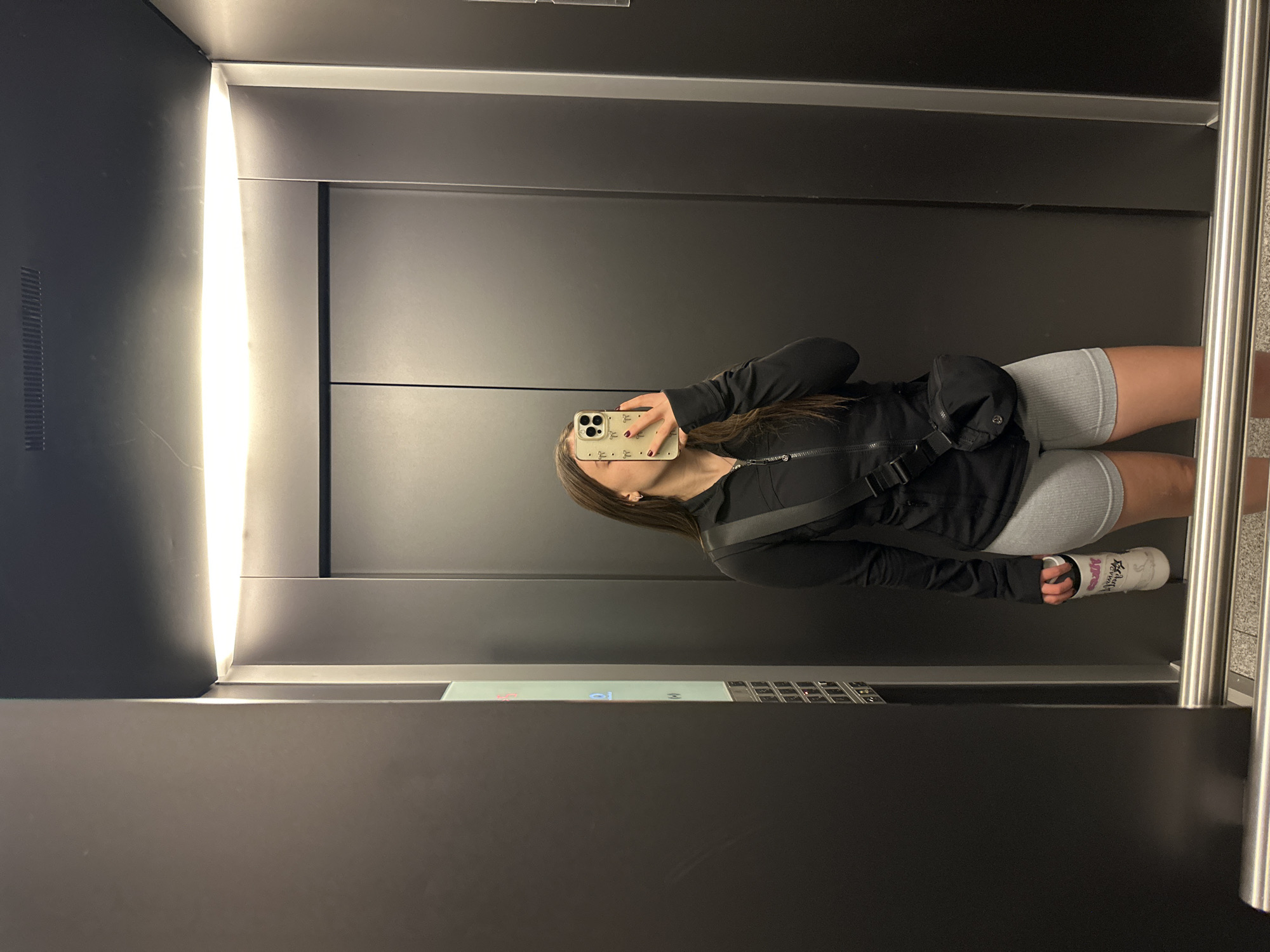 A woman is taking a selfie in an elevator. She is wearing a black zip-up jacket, grey shorts, and a white bandage on her left wrist. She is holding a phone with a floral-patterned case. The elevator has grey walls and a metal control panel. It is illuminated by a bright light panel. It is not possible to verify the location.