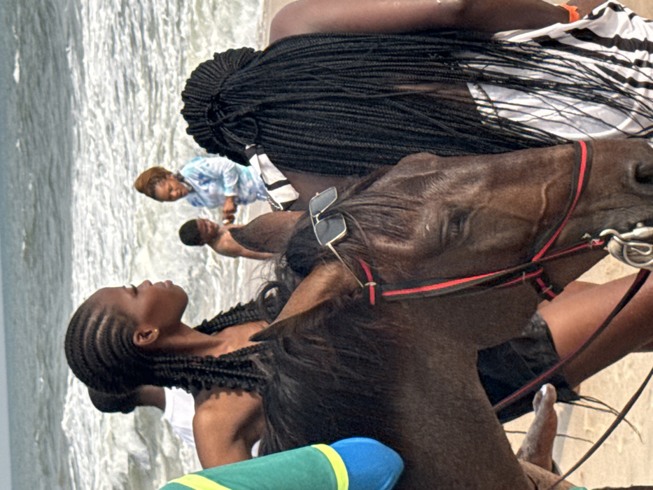 A group is on a beach. A woman with braided hair looks upwards while riding a dark-brown horse. Another woman with braids stands nearby. In the background, a person in a blue-and-white patterned shirt walks along the water. Another person is laying in the shallow water. The horse has sunglasses and a red bridle.