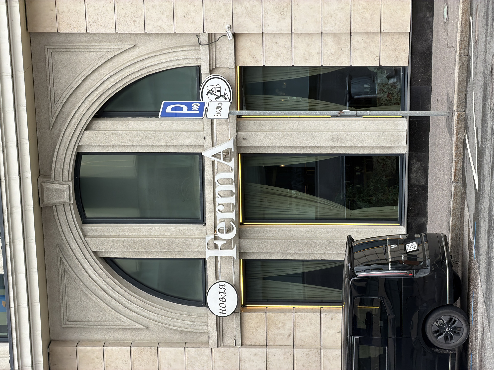 A vertically oriented photograph shows the exterior of a building in Saint Petersburg, Russia, displaying the "Forma" restaurant. The facade is clad in light-colored stone or tile, with distinctive arched windows featuring black frames. Next to the window is the restaurant name “Forma” displayed vertically. To the left of the restaurant’s name there are signs and symbols with a parking symbol, and other symbols and lettering in Russian. A black vehicle is parked on the street beside the building, partially obscuring the lower portion of the restaurant's exterior. The sidewalk or pavement is visible to the right of the vehicle.