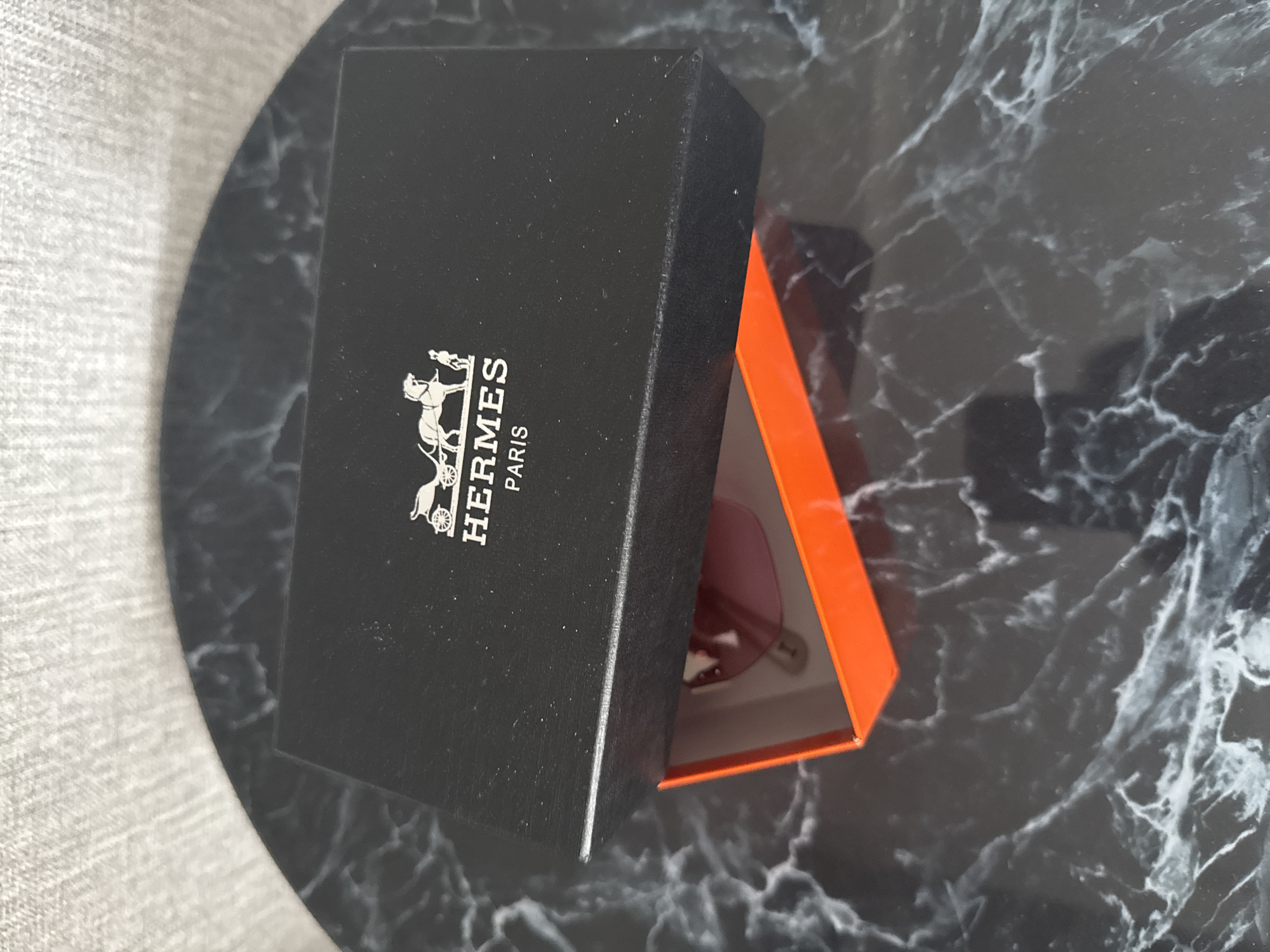 A black Hermès box is open, revealing a pair of pink sunglasses inside. The box is placed on a dark marble surface. The background is a textured, light-colored fabric. The image was taken in the city of Soloneshnoye, Russia.