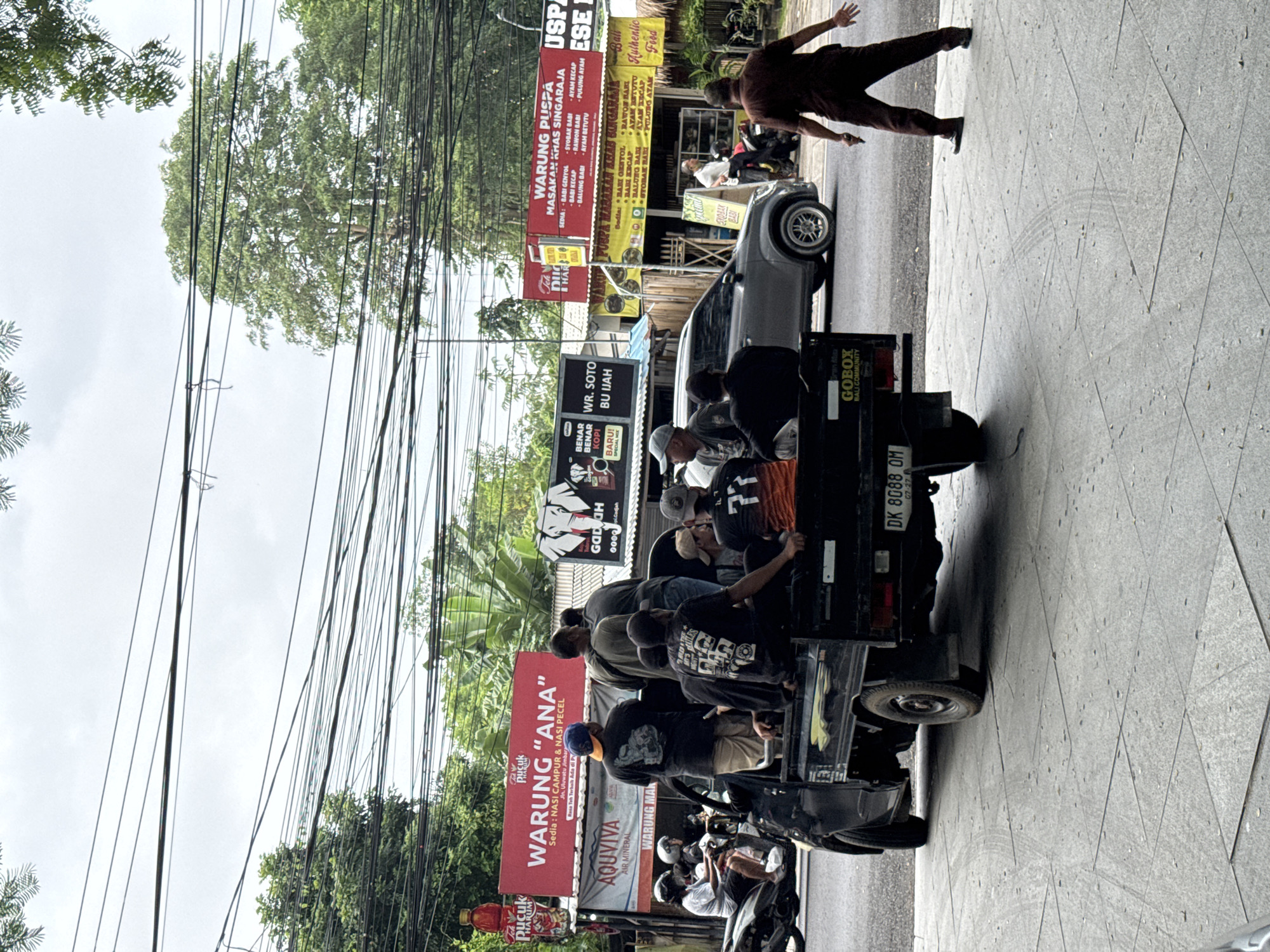The image captures a street scene in Kuta Selatan, Indonesia. A black utility vehicle is positioned on the right side of the frame, its truck bed filled with people. Other vehicles, including cars and motorbikes, are parked nearby. Power lines are visible overhead, crisscrossing the sky. Various signs advertising local businesses, such as "WARUNG PUSPA" and "WARUNG 'ANA'," line the street. A man in a brown outfit appears to be walking on the sidewalk with his arms extended. The street is paved with a textured gray surface.