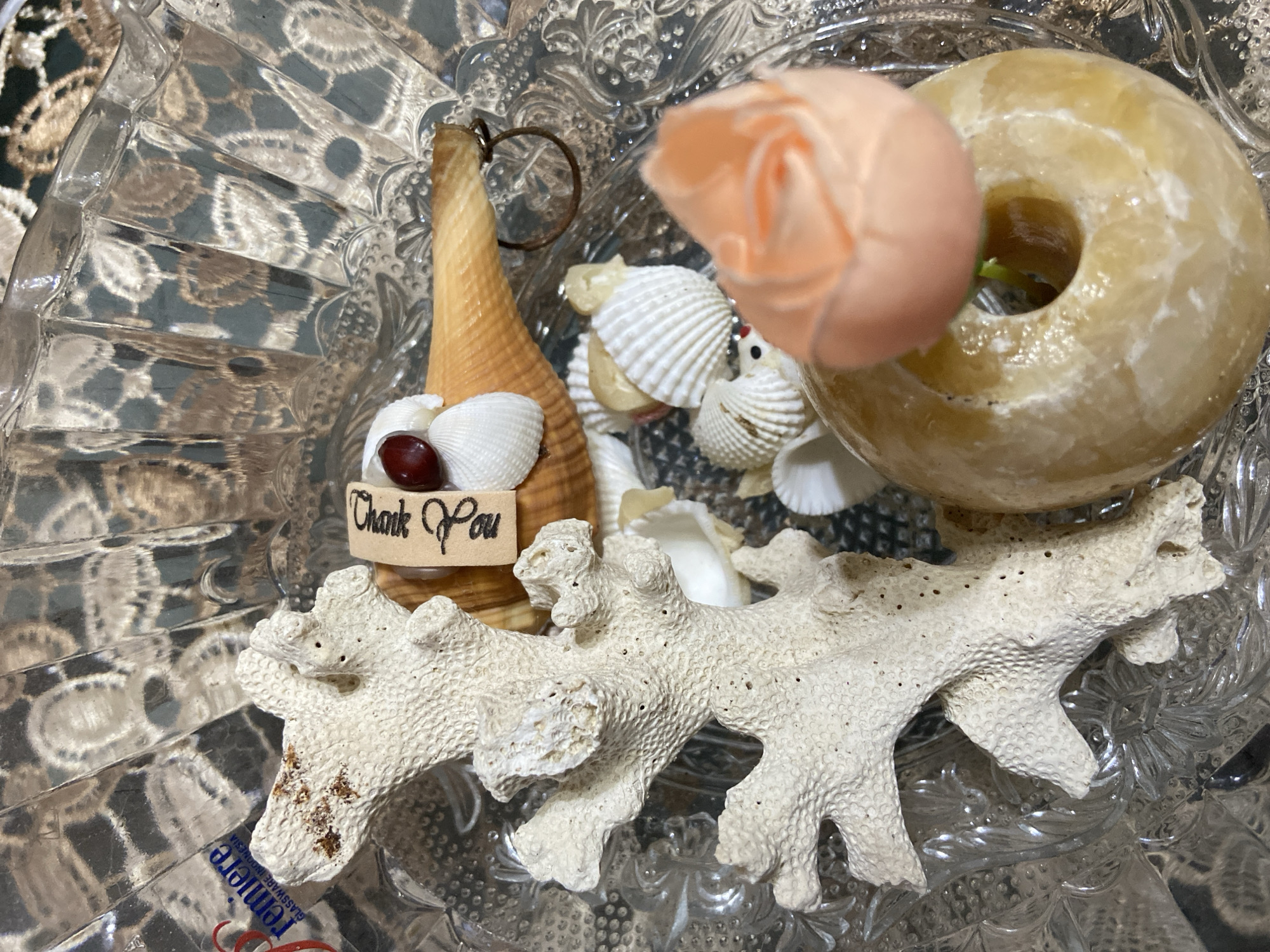In the city of Margorejo, Indonesia, a close-up, overhead shot showcases a decorative arrangement within a clear, faceted glass bowl. The focal point is a collection of beach-themed items: a piece of white coral, several small seashells, a larger shell, and a ring. A small, dark red bead is affixed to a cluster of white shells. Attached to the larger shell is a small, wooden tag with the handwritten words "Thank You". Partially visible in the bottom right is a donut-shaped, yellowish object topped with a pale orange, rose-shaped ornament. The background of the bowl reveals reflections of a patterned fabric or surface.