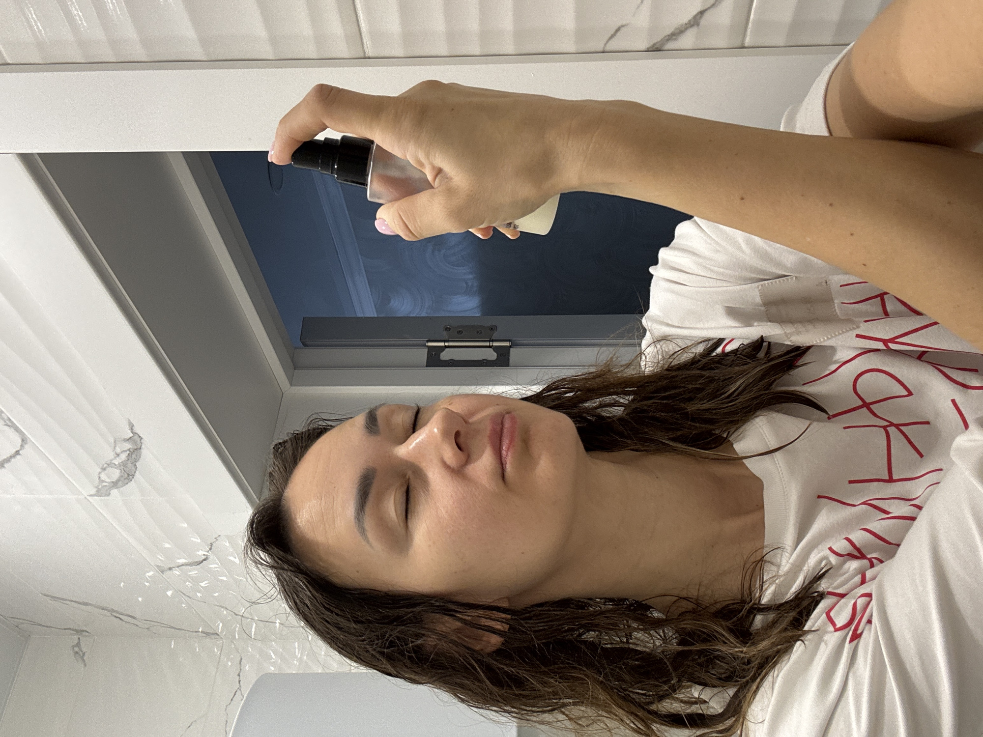 A person with dark brown hair, eyes closed, is shown holding a small spray bottle in front of their face. They are wearing a white t-shirt with a red design. The background includes white marble tile, a white cabinet, and a dark reflection. The setting appears to be a bathroom. It is not possible to confirm a location in Sochi, Russia based solely on the visual information provided.