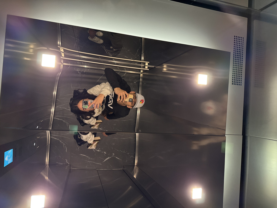 A couple takes a selfie in a modern elevator with reflective walls and ceiling. The woman has long dark hair and is wearing a light-colored sweatshirt, while the man has a baseball cap and a dark sweatshirt. The elevator has a sleek, dark marble floor and minimalist lighting fixtures. A control panel is visible on the left, and ventilation grates are on the right. Their reflections appear on the ceiling and floor of the elevator.