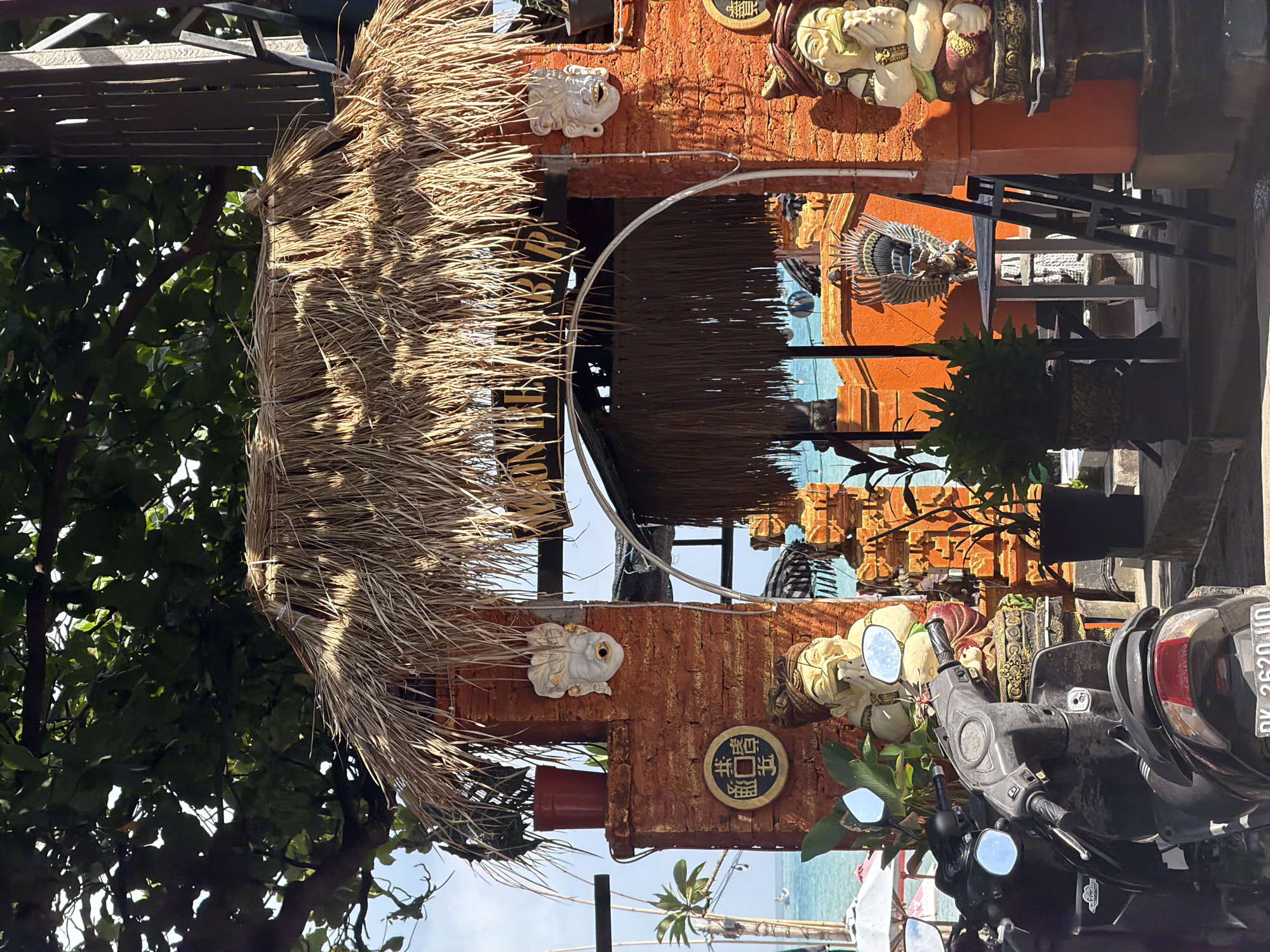 A scene unfolds in Kuta, Indonesia, featuring an outdoor establishment with a thatched roof entrance adorned with traditional Balinese artistry. The entrance is framed by brick pillars decorated with masks and statues, including a seated Buddha-like figure and a winged creature. Scooters are parked in the foreground, obscuring part of the entrance. Beyond the entrance, a glimpse of the ocean and beach umbrellas suggests a coastal location. The overall ambiance is that of a tropical, leisurely setting.