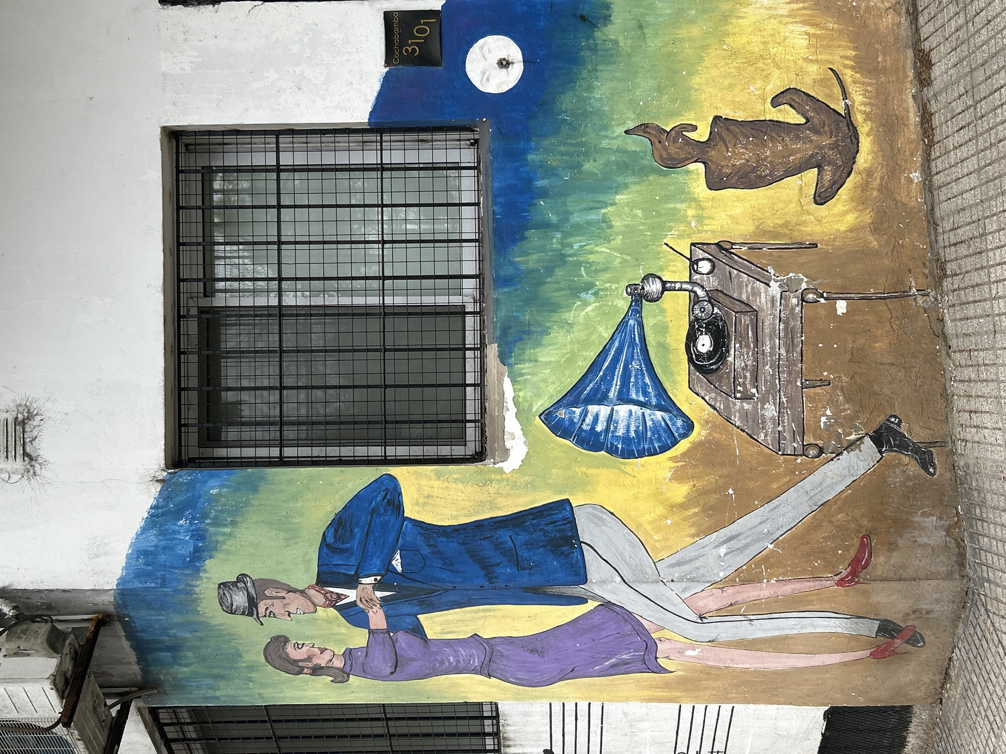 This is a photograph of a mural painted on the exterior wall of a building in Buenos Aires, Argentina. The mural depicts a couple dancing in a style that suggests tango, set against a background of green and yellow, with a blue wash at the top. The man is wearing a blue suit, white shirt, and a fedora. The woman is in a purple dress. To the right of the couple, there is a vintage record player with a blue horn, placed on a wooden table. Further to the right, on the yellow background, is a stylized brown figure that could represent a flame or an abstract sculpture. Above the mural, a window with a black metal security grate is visible. On the upper right section of the wall, a small black plaque reads "Cochabamba 3101". A circular white object with a face-like design is painted to the left of the plaque. The mural is weathered, with some cracks and peeling paint visible on the wall. The lower part of the image shows a paved sidewalk with a grid pattern.