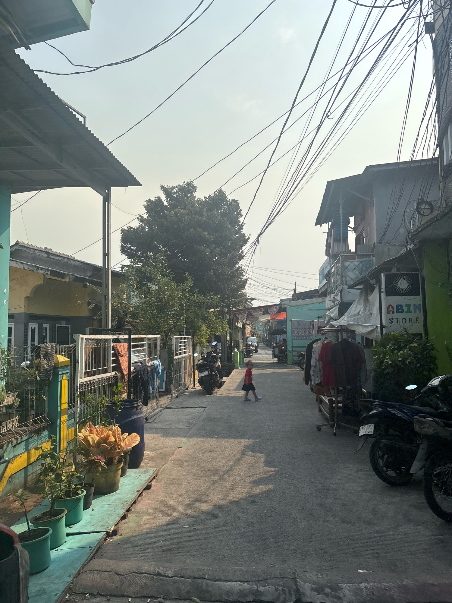 The image depicts a narrow, paved alleyway in a densely populated urban environment, likely a residential area within North Jakarta, Indonesia. The alley is flanked by buildings on both sides, constructed with a mix of corrugated metal and painted plaster. Overhead, a dense network of electrical wires crisscrosses the sky.
On the left side of the alley, a building with a yellow facade is visible, featuring a fenced-off area with several potted plants. A metal gate with laundry hanging on it separates the property from the alley. Further down the alley on the left, a large tree provides shade.
On the right side of the alley, a series of buildings house small businesses. One establishment, identified by a sign reading "ABIN STORE," is clearly visible. Clothes are displayed on racks outside another shop. Several motorcycles are parked along the right side of the alley, suggesting a common mode of transportation.
A single child, wearing a red shirt and dark shorts, is walking down the center of the alley away from the camera. The sun is casting long shadows, indicating it is likely late afternoon. The overall impression is one of a vibrant, lived-in urban space, common in many Indonesian cities.